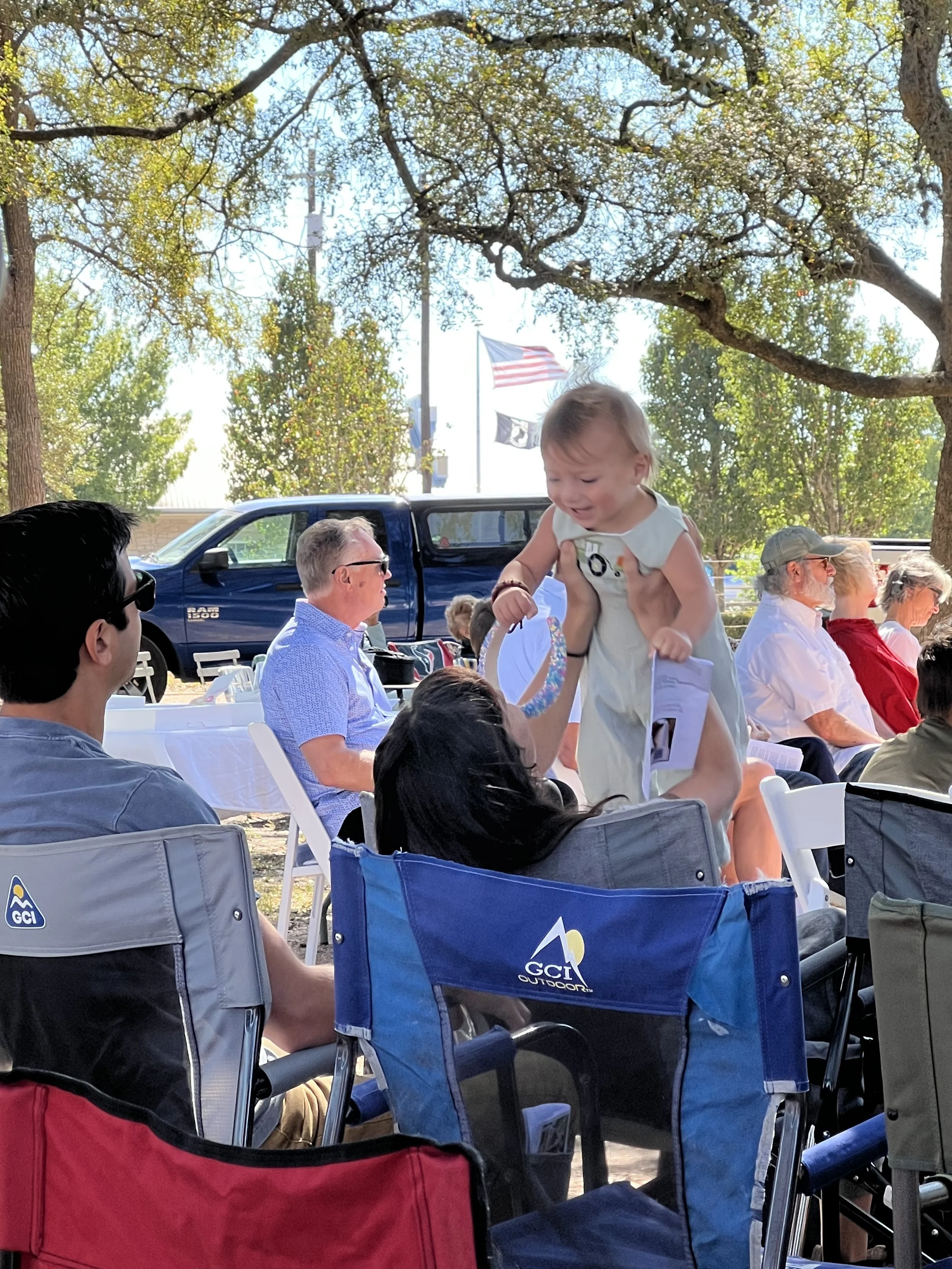 A group of people gathered outdoors sitting on chairs, with some wearing sunglasses and casual clothing. A young child being lifted in the air by an adult is visible in the center. Behind the group, SUVs and American flags are seen under a large tree