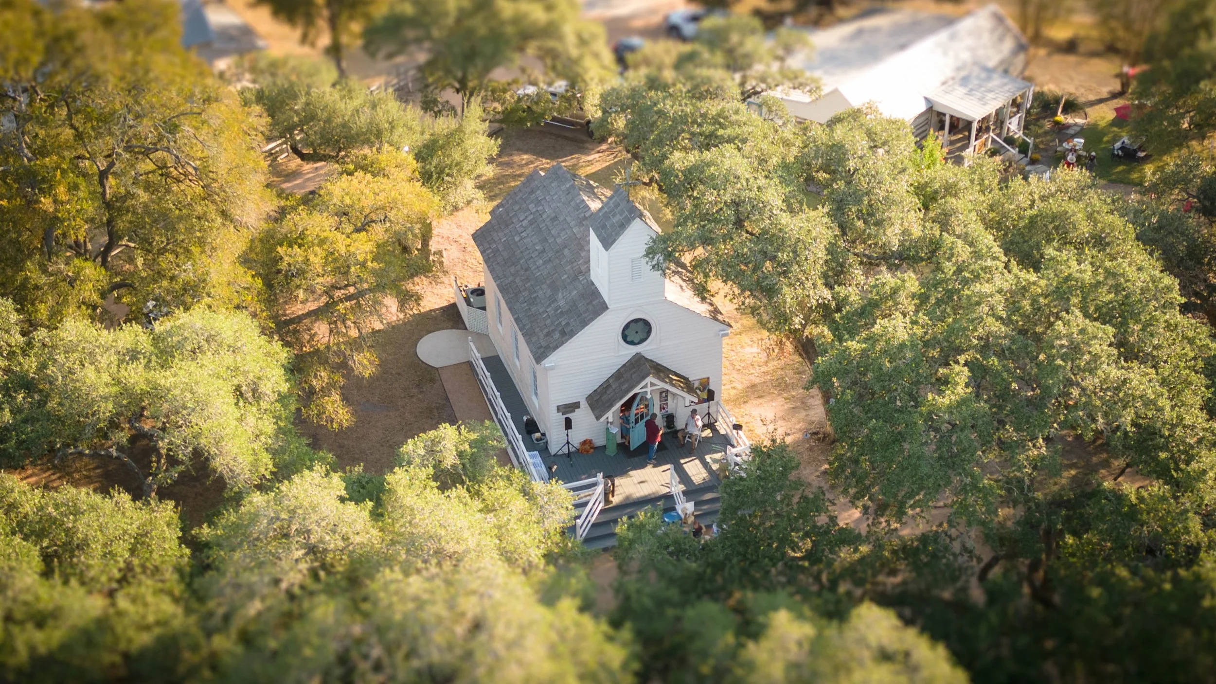 A small white church with a wooden deck in front, surrounded by trees with green leaves, seen from above during golden hour.