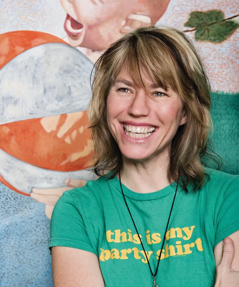 A woman smiling with light brown, shoulder-length hair, wearing a green T-shirt that says "this is my party shirt" in yellow text, standing in front of a colorful mural.