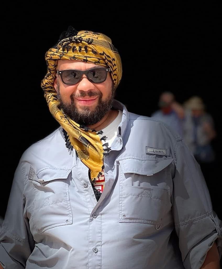 A man wearing sunglasses, a yellow and black patterned headscarf, a white button-up shirt, and smiling outdoors with blurred background of other people.