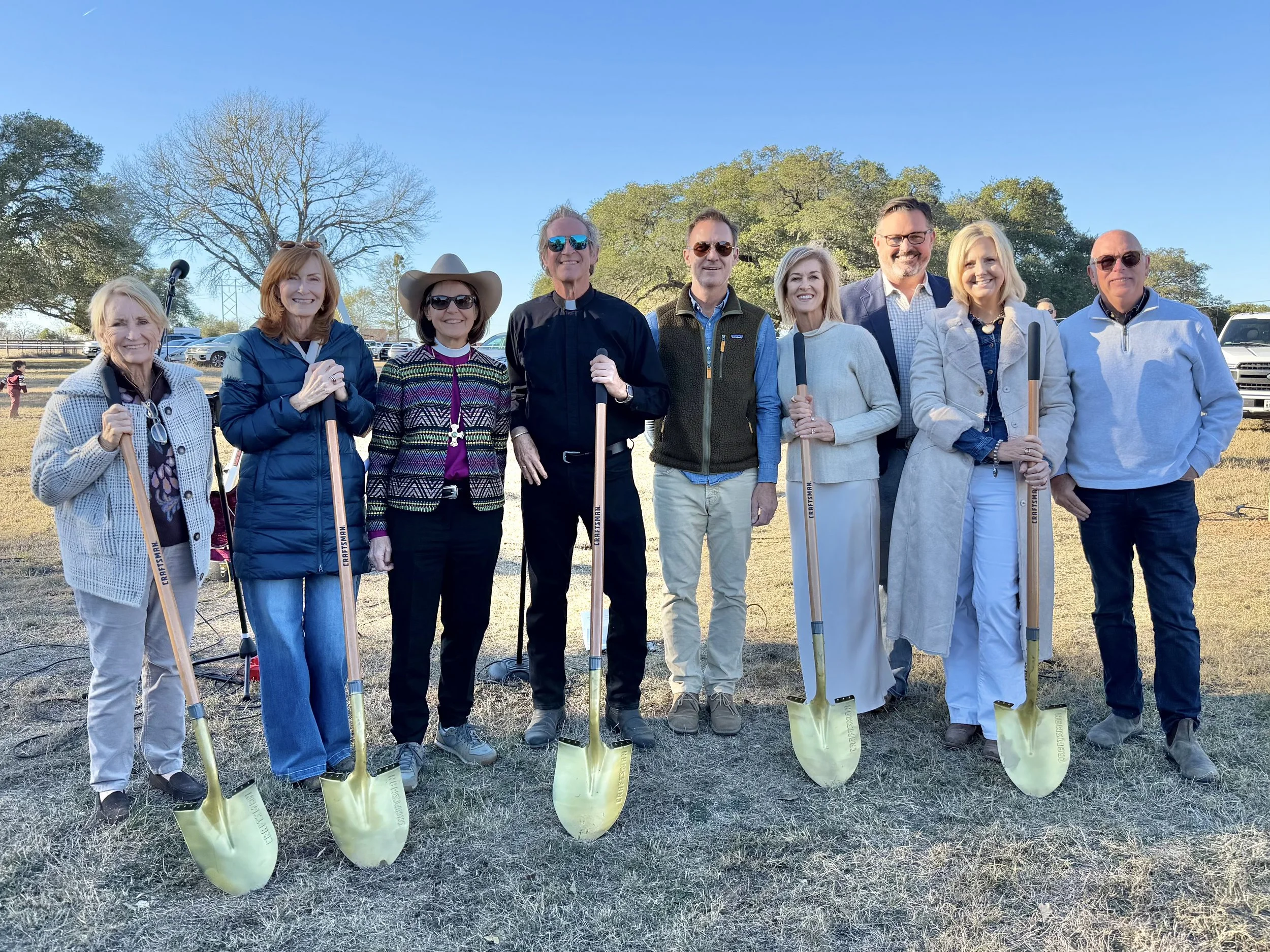 A group of people standing outdoors in a park or open field, holding shovels planted into the ground, smiling for a photo, with trees in the background.