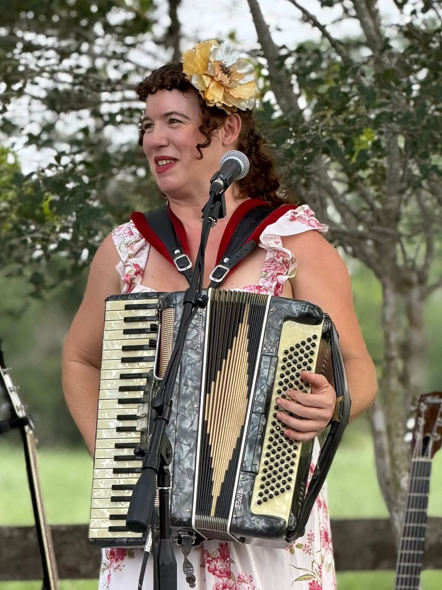 Woman wearing a floral dress and a large flower hair accessory, playing an accordion outdoors, with trees in the background.