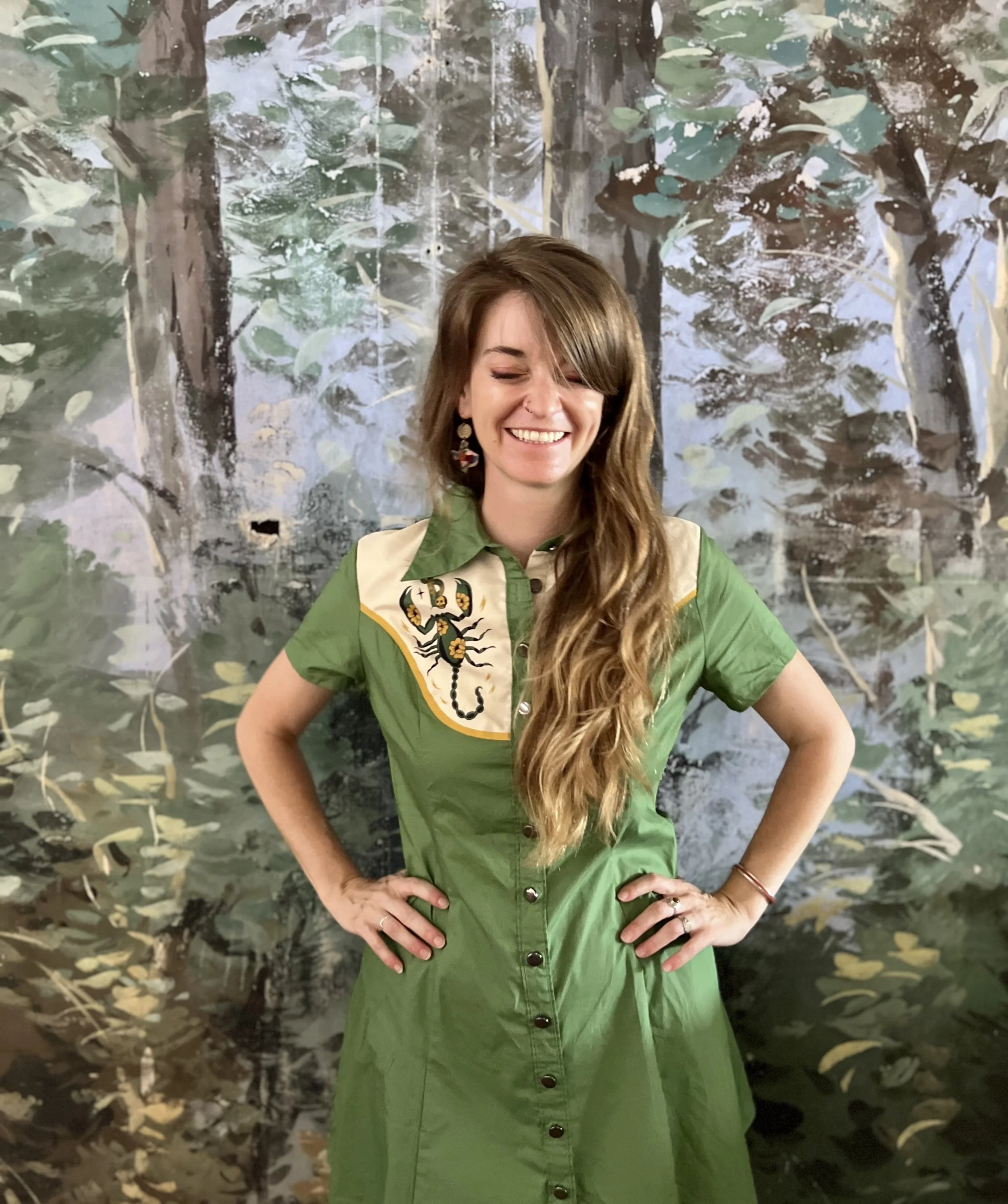 A woman in a green dress with a beige sash with a floral and scorpion design. She is smiling with hands on her hips, standing in front of a painted forest background.