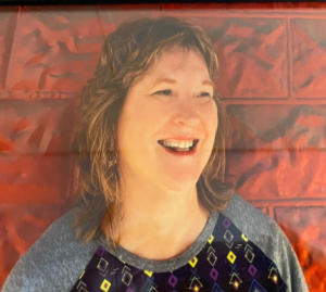 A woman with shoulder-length curly hair smiling against a red textured wall.