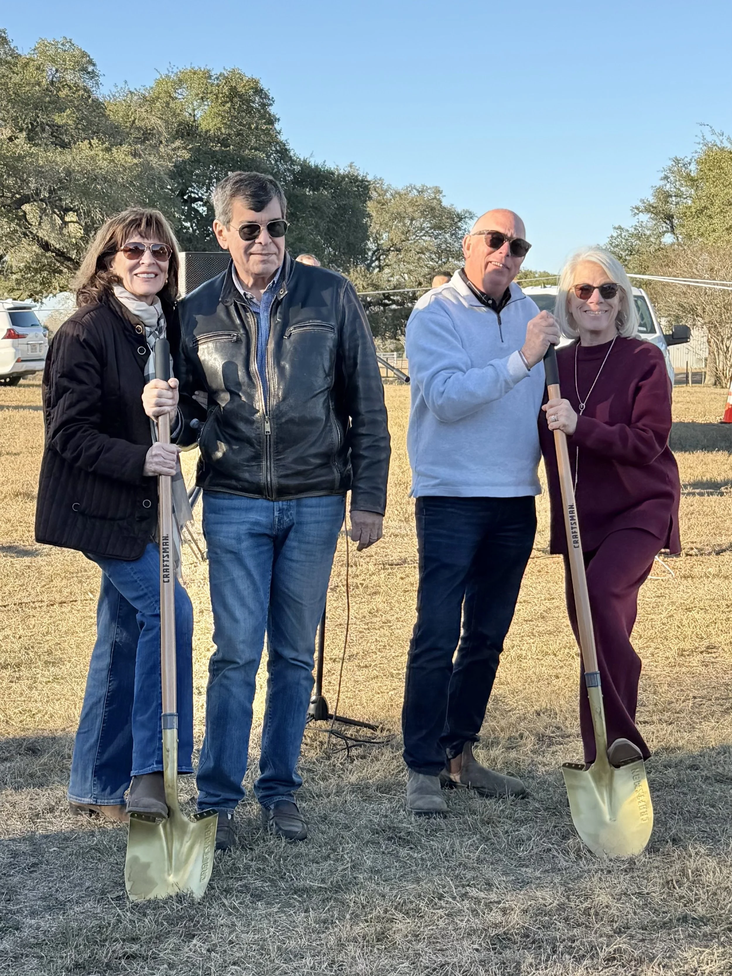 Four adults standing outdoors on grass, holding shovels, smiling, with trees and parked cars in the background.