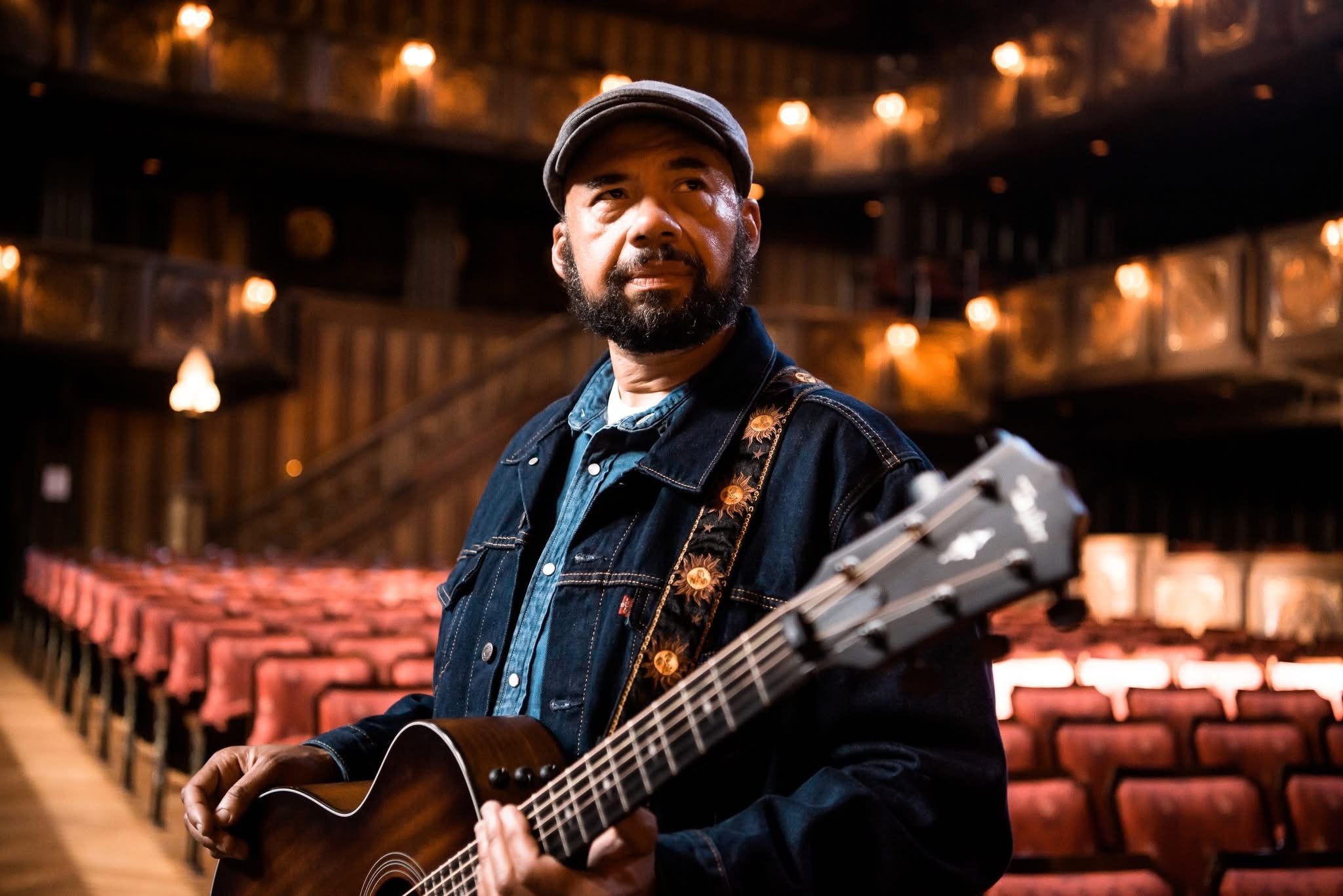 A man with a beard and cap playing an acoustic guitar in an empty theater with red seats and warm lighting.