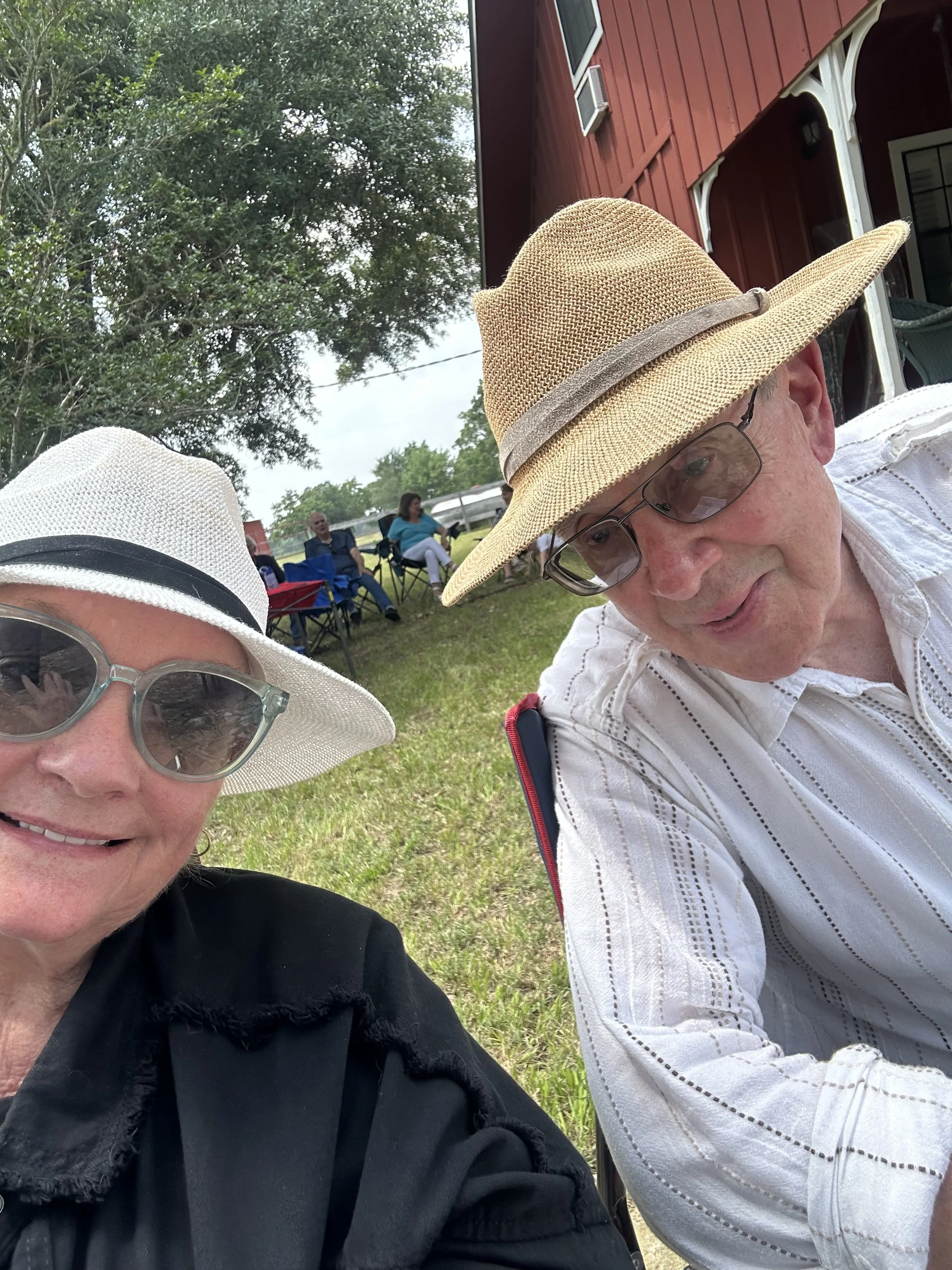 A close-up selfie of two seniors outdoors, wearing sun hats and glasses, with a background of trees, a red building, and other people sitting on lawn chairs.