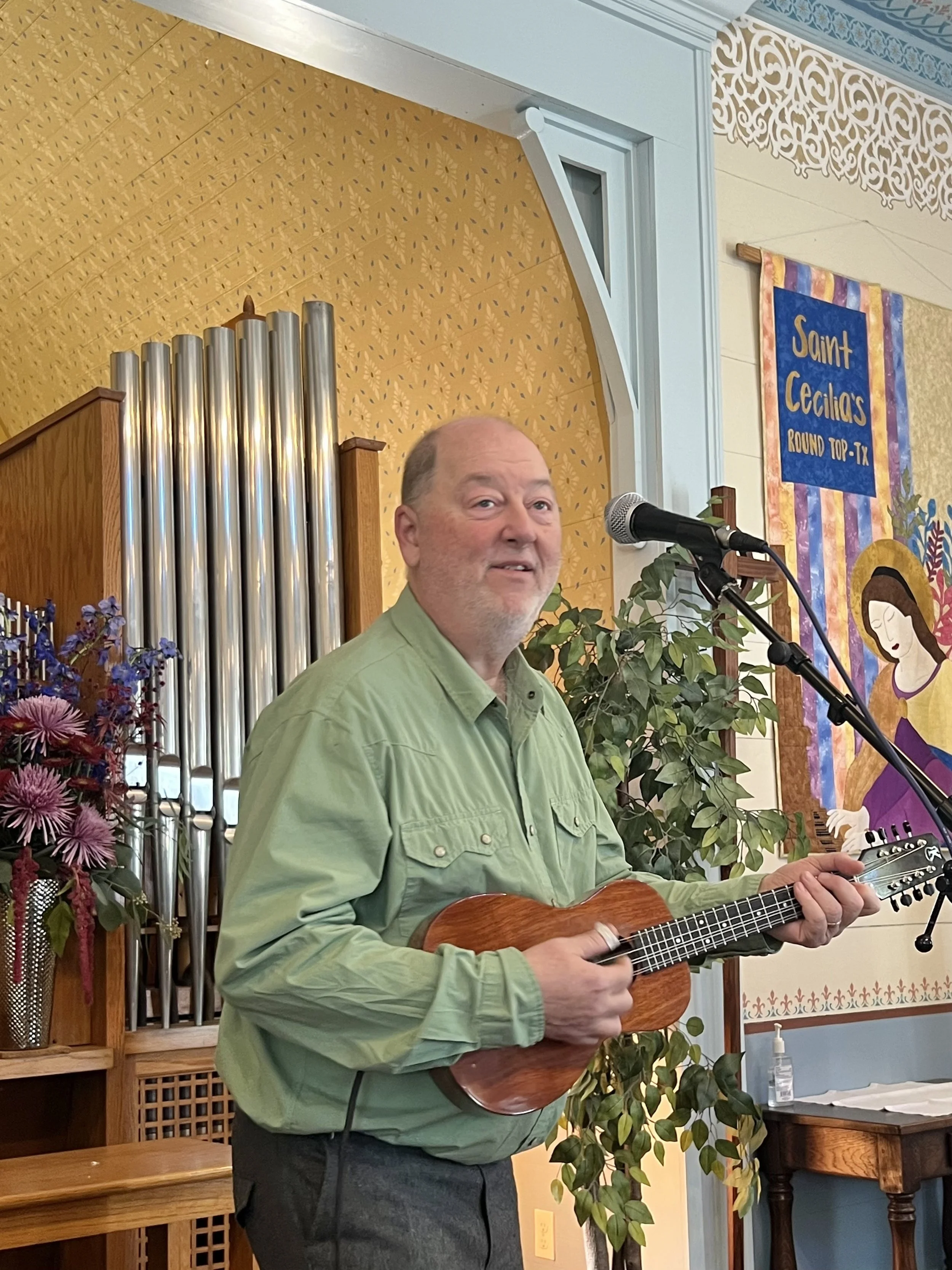 A man in a green shirt playing a ukulele and singing at a small indoor venue, with a microphone and musical pipes behind him, decorated with flowers and banners.