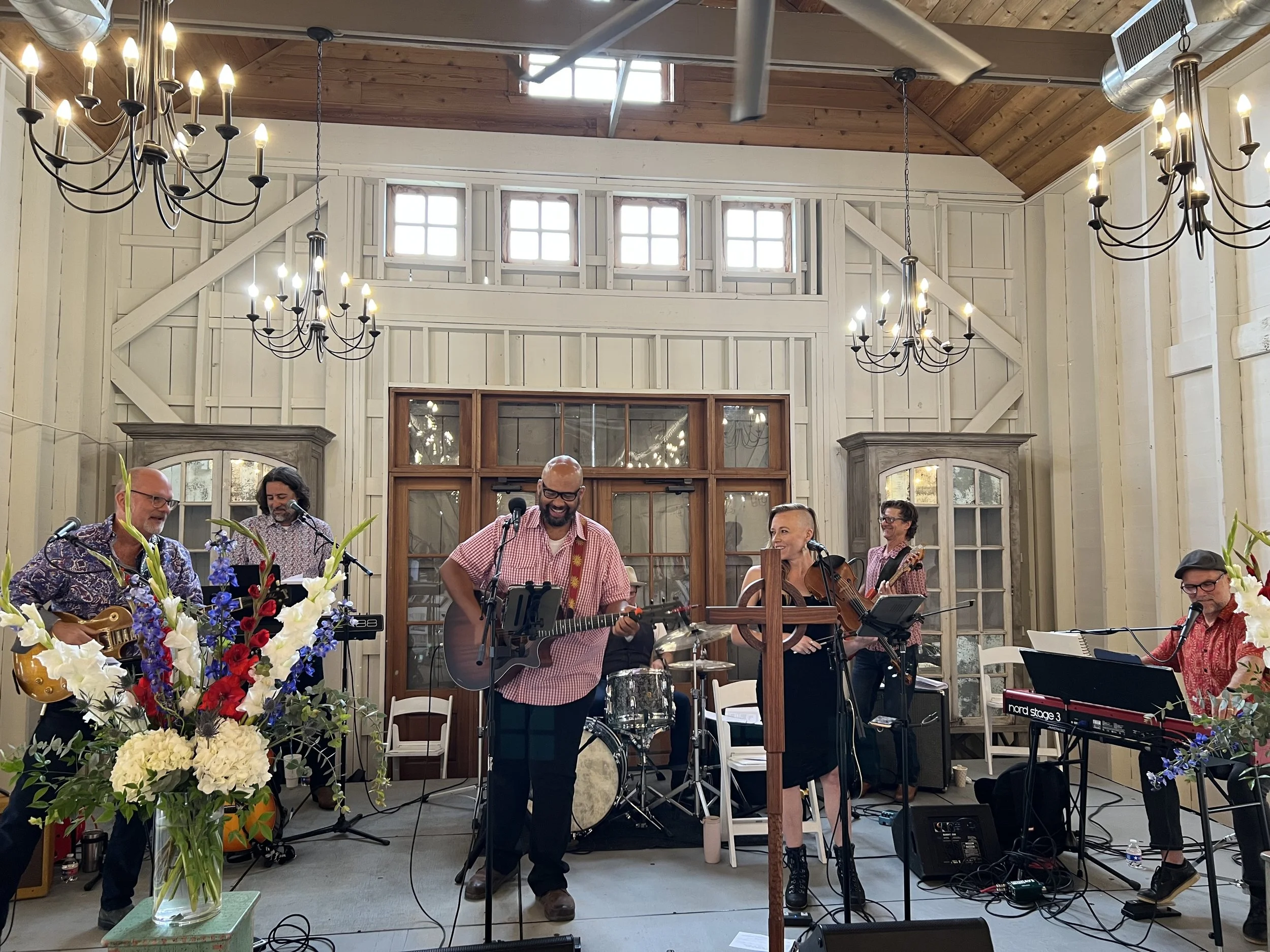 A band performing in a rustic indoor space with chandeliers, wooden walls, and large windows, featuring musicians playing guitars, keyboard, drums, and violin, with a large floral arrangement in the foreground.