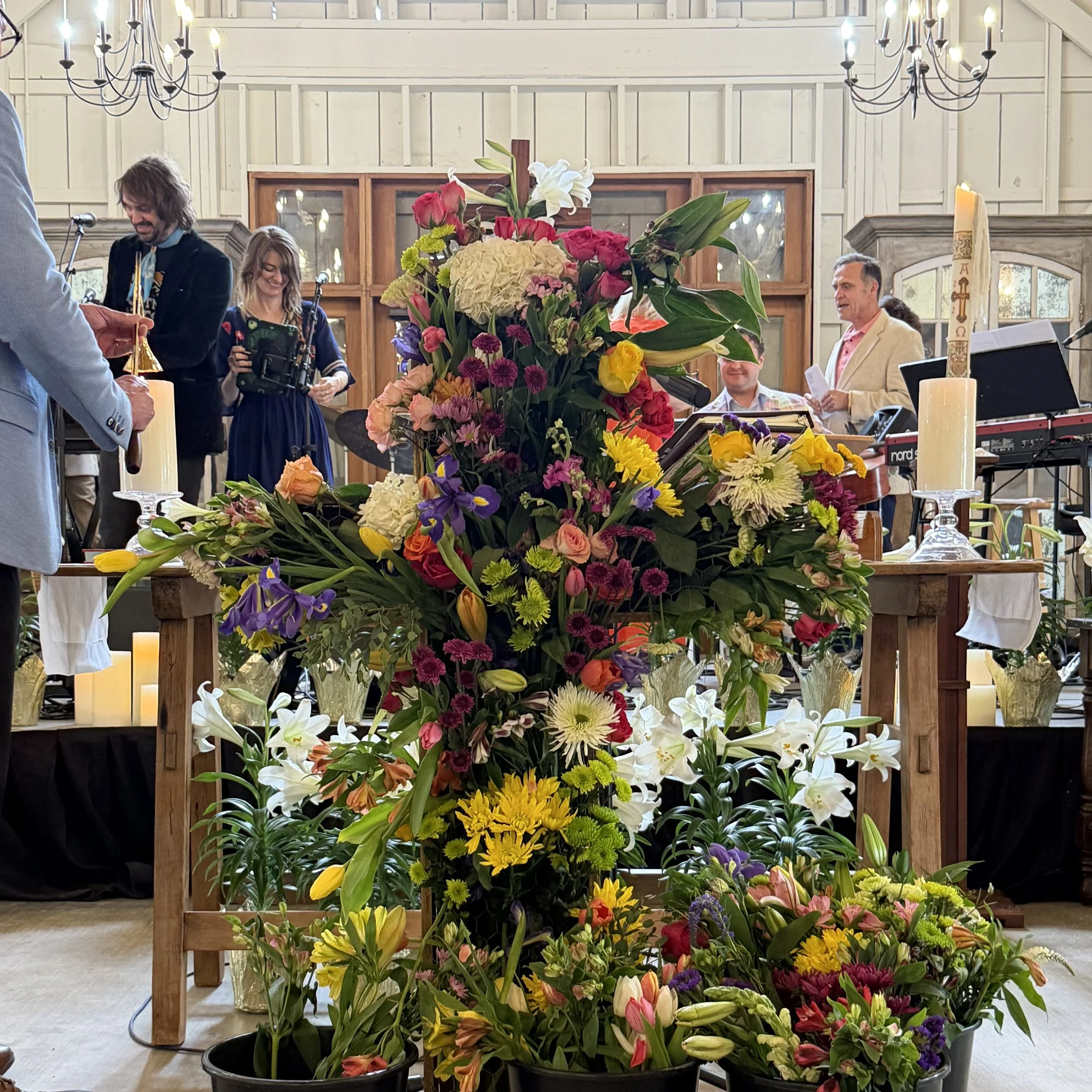 A large arrangement of colorful flowers on a table inside a building, with people gathered around, some holding papers or microphones, and candles and chandeliers in the background.