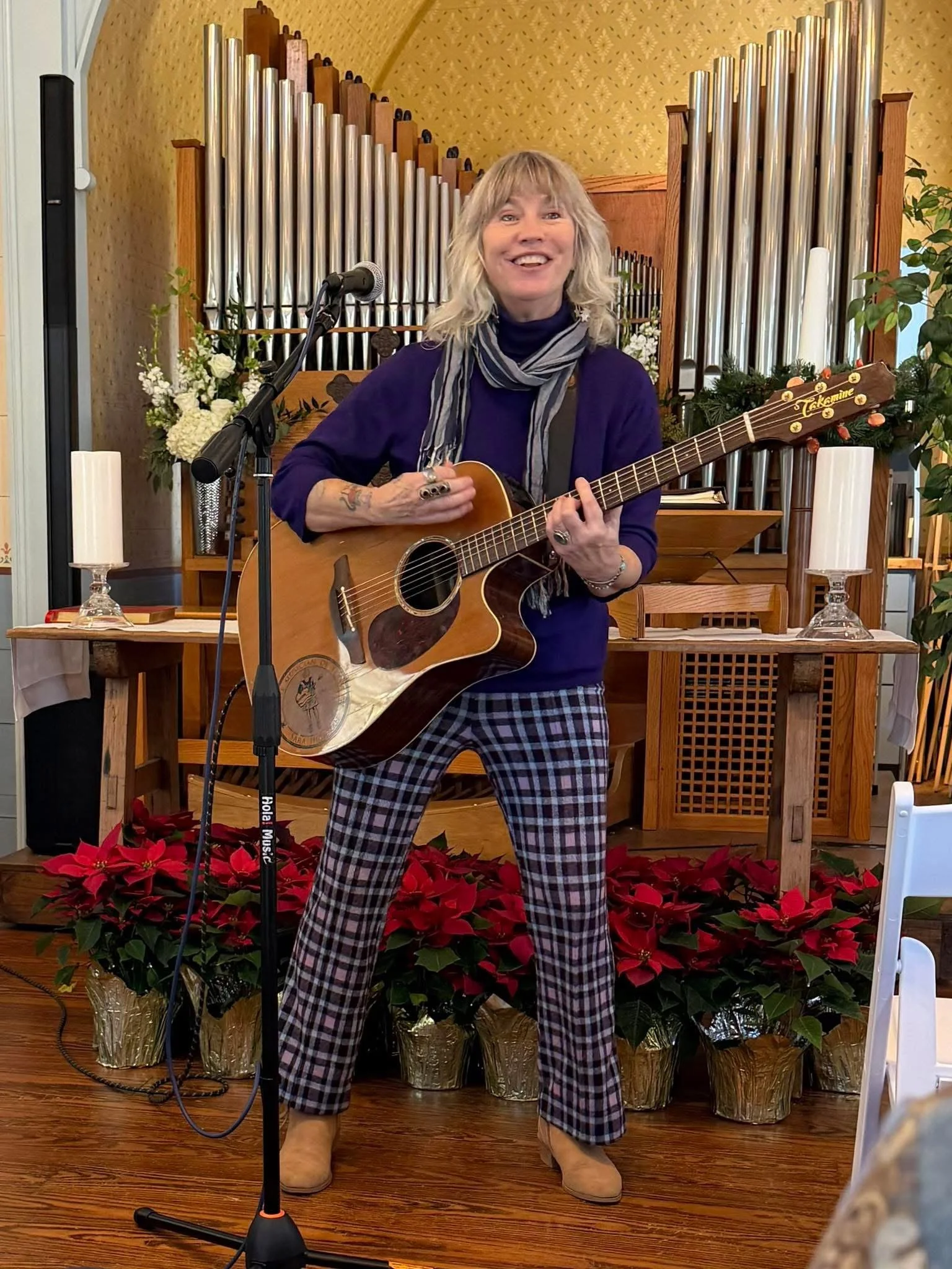 Person with curly blond hair playing guitar and singing into a microphone indoors, with a pipe organ in the background and poinsettia plants in front.