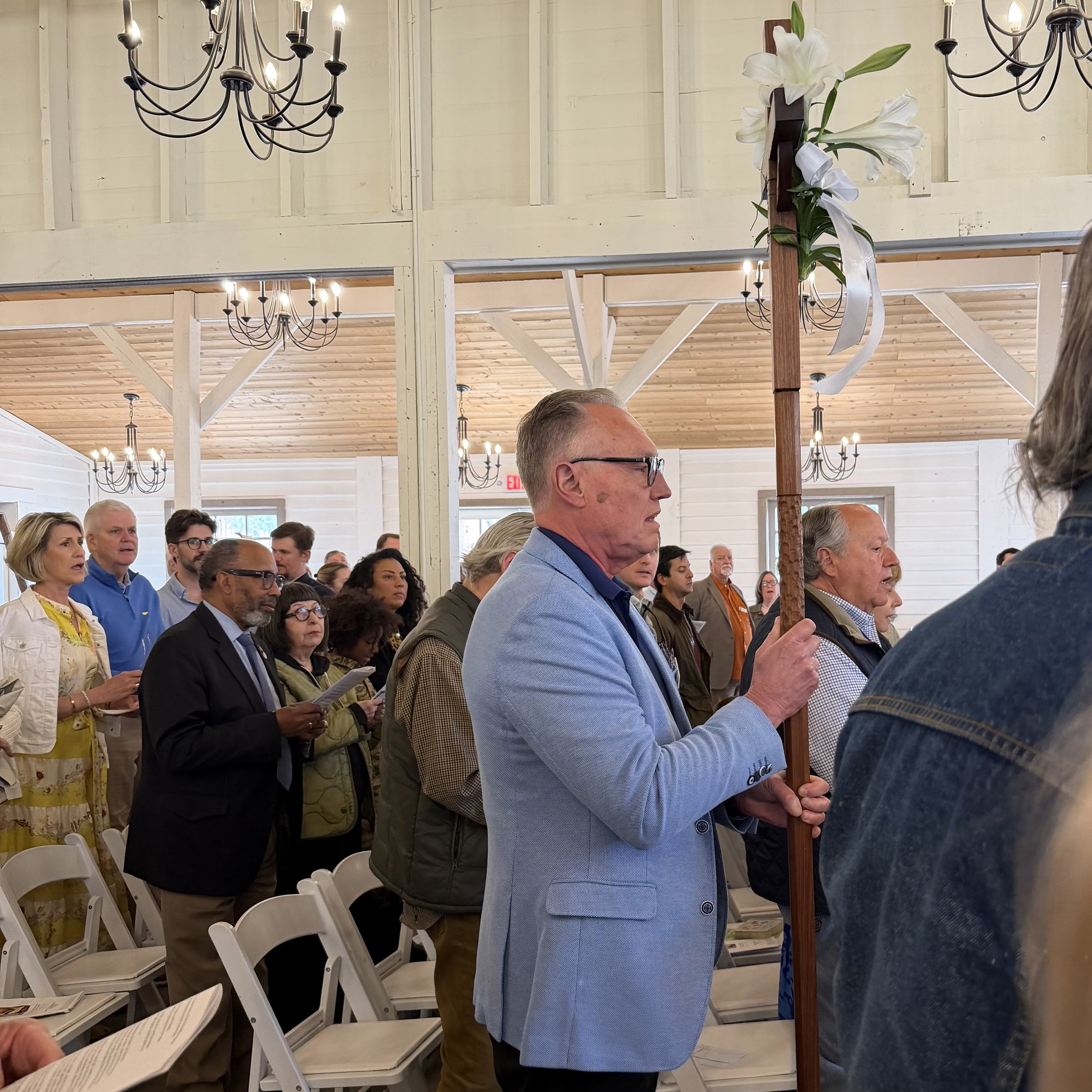 People gathered indoors, dressed in formal and semi-formal attire, standing and singing during an event with chandeliers and a floral arrangement in the background.