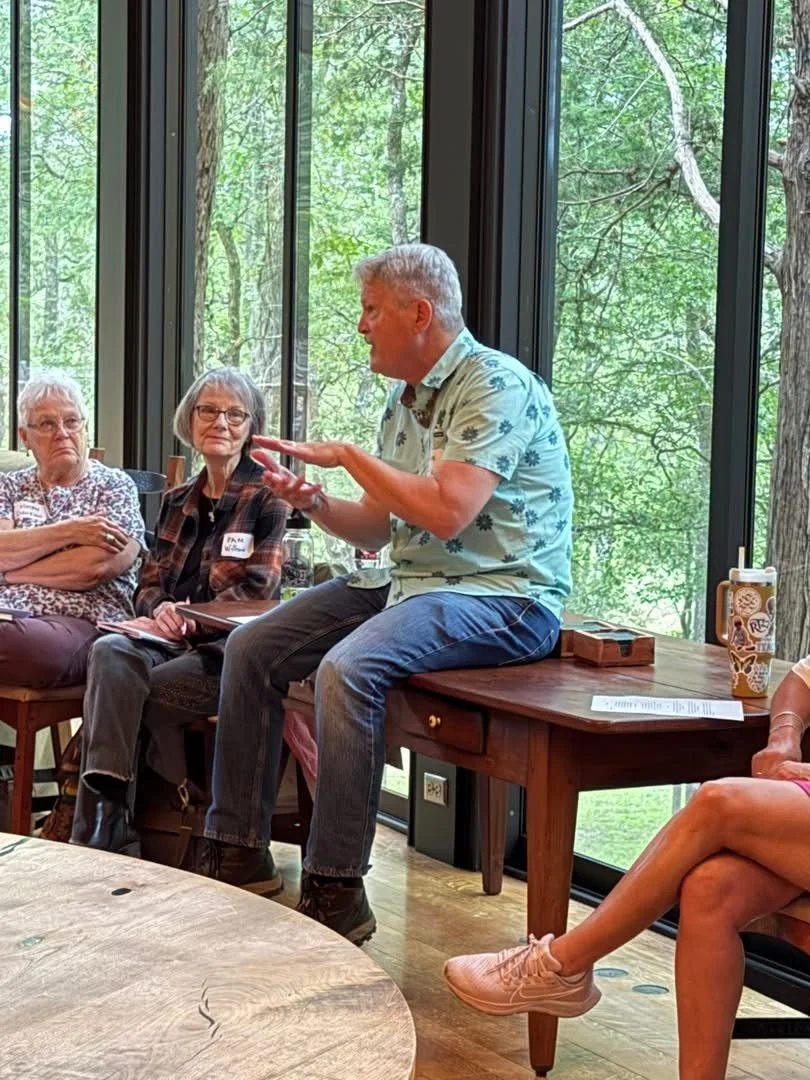 Man in floral shirt speaking to a group in a room with large windows and a view of trees outside.