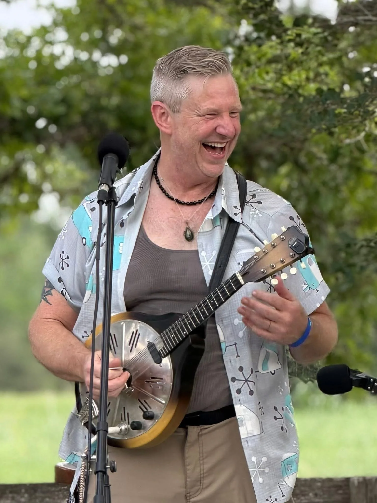 A middle-aged man with short gray hair is laughing while playing a metallic resonator guitar outdoors. He is wearing a patterned short-sleeve shirt over a gray T-shirt, a beaded necklace, and a wristband. There is a microphone on a stand in front of 