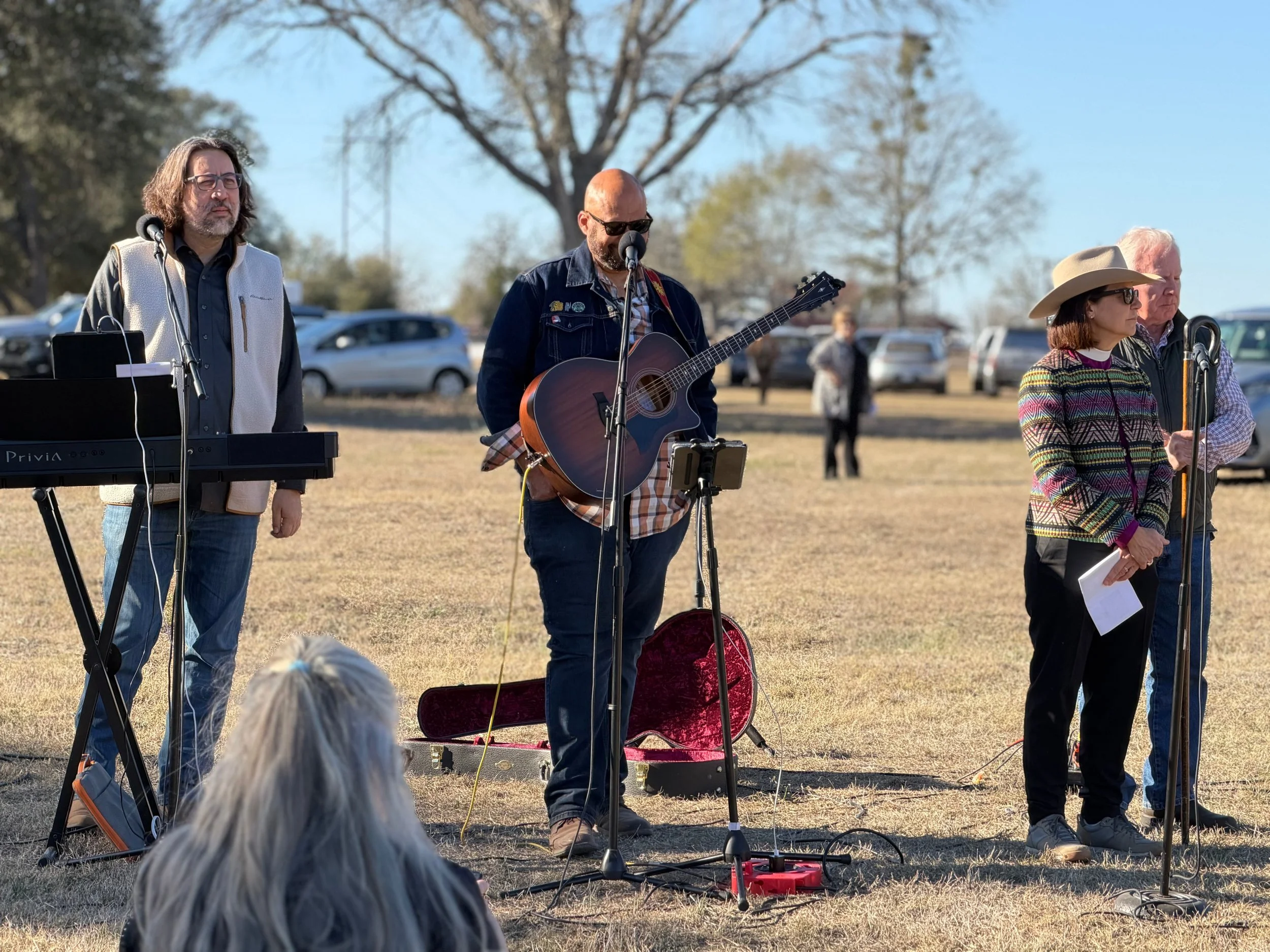 Group of four musicians performing outdoors, with one playing keyboard, one playing guitar, and others standing nearby, in a park-like setting with trees and parked cars.