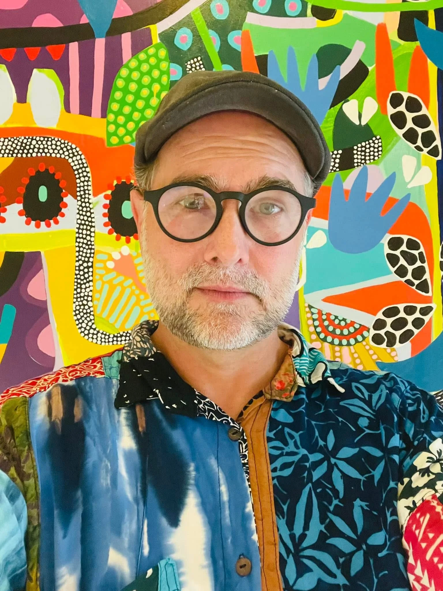 A man with light skin, glasses, and a gray beard wearing a colorful patterned shirt and a hat, standing in front of a vibrant, abstract, multicolored artwork featuring various shapes and patterns.