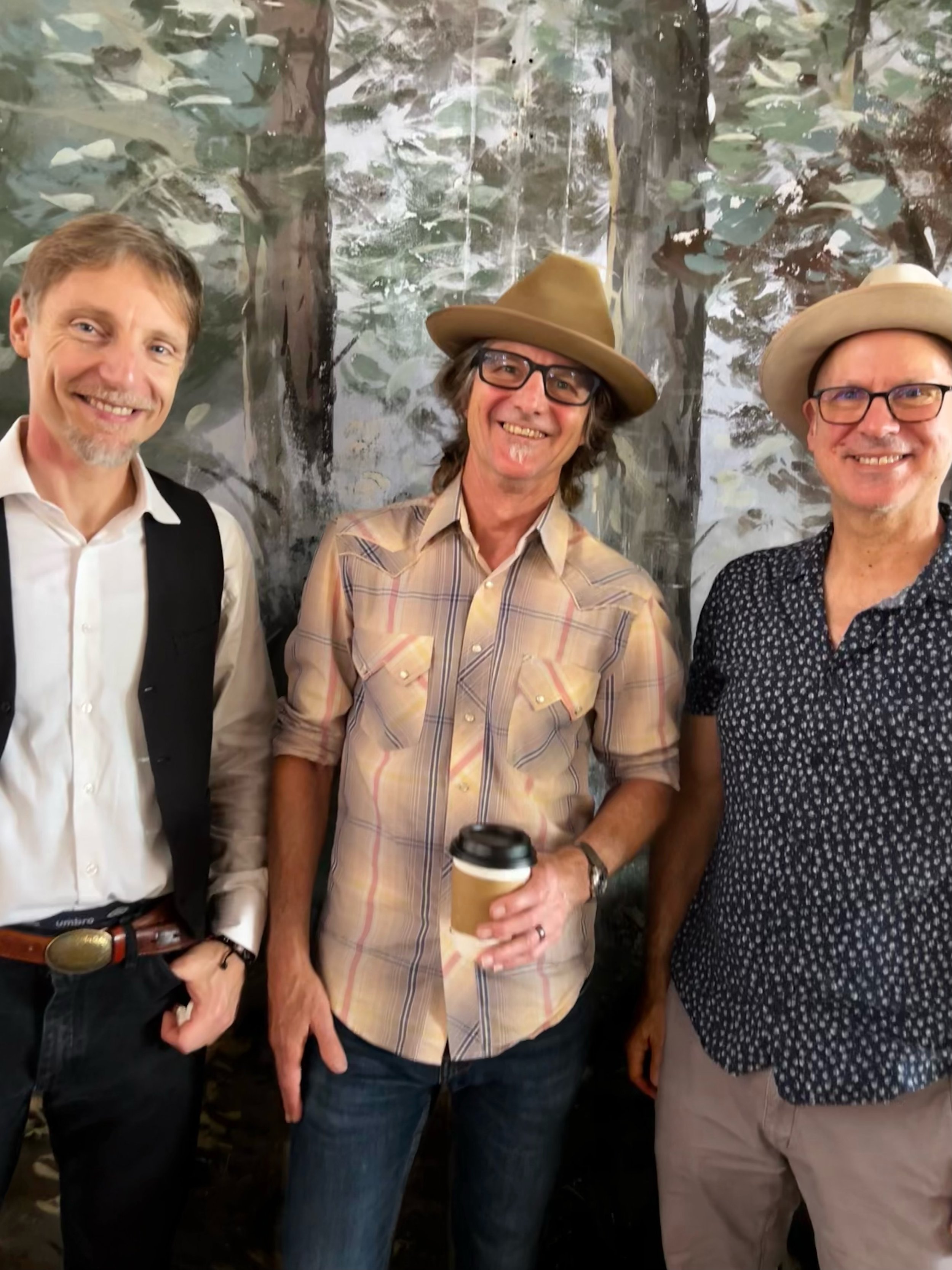 Three men standing together smiling in front of a painted forest backdrop. Two are wearing hats, and one is holding a cup of coffee.