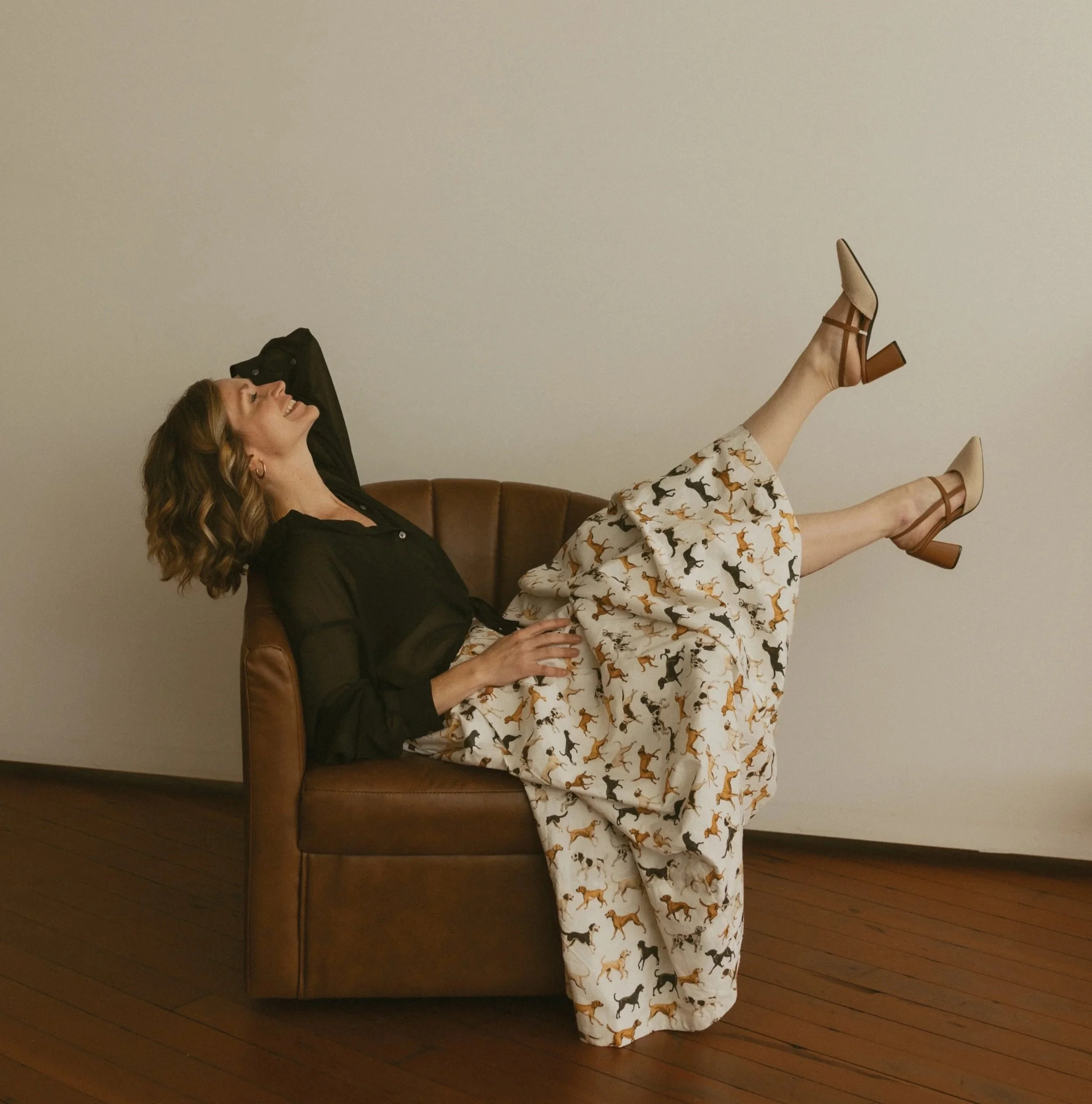 A woman with curly hair lying on a brown leather couch, smiling and looking up, with her arms behind her head. She is wearing a black top and patterned wide-leg pants with animals printed on them. Her legs are raised and crossed at the ankles, wearing beige heels. The background features a plain, light-colored wall and wooden floor.