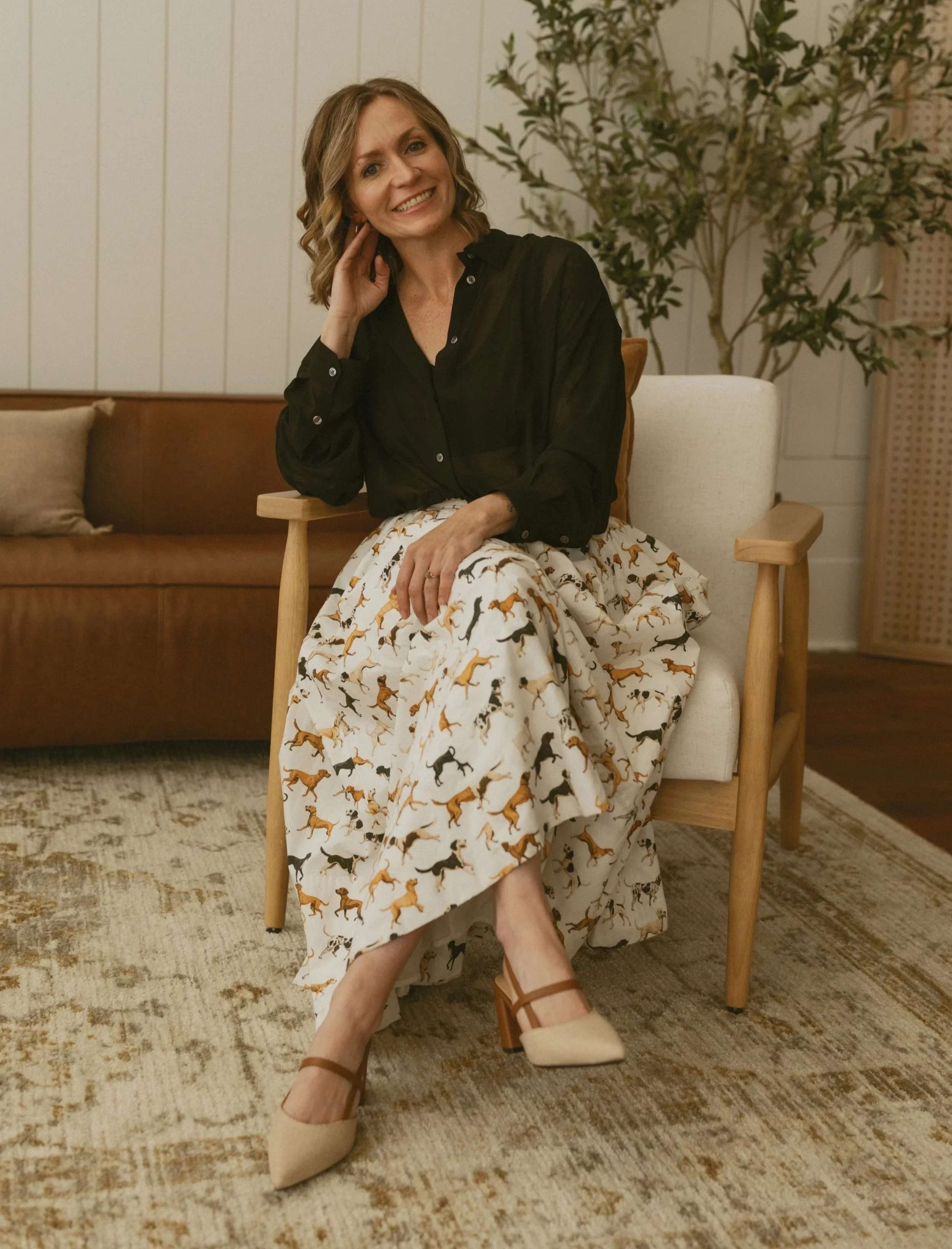 A woman with shoulder-length hair, wearing a black sheer blouse and a long skirt with a dachshund dog pattern, sitting on a wooden chair in a living room with a beige and brown rug, a beige armchair, and a background of white-paneled walls and a large green plant.