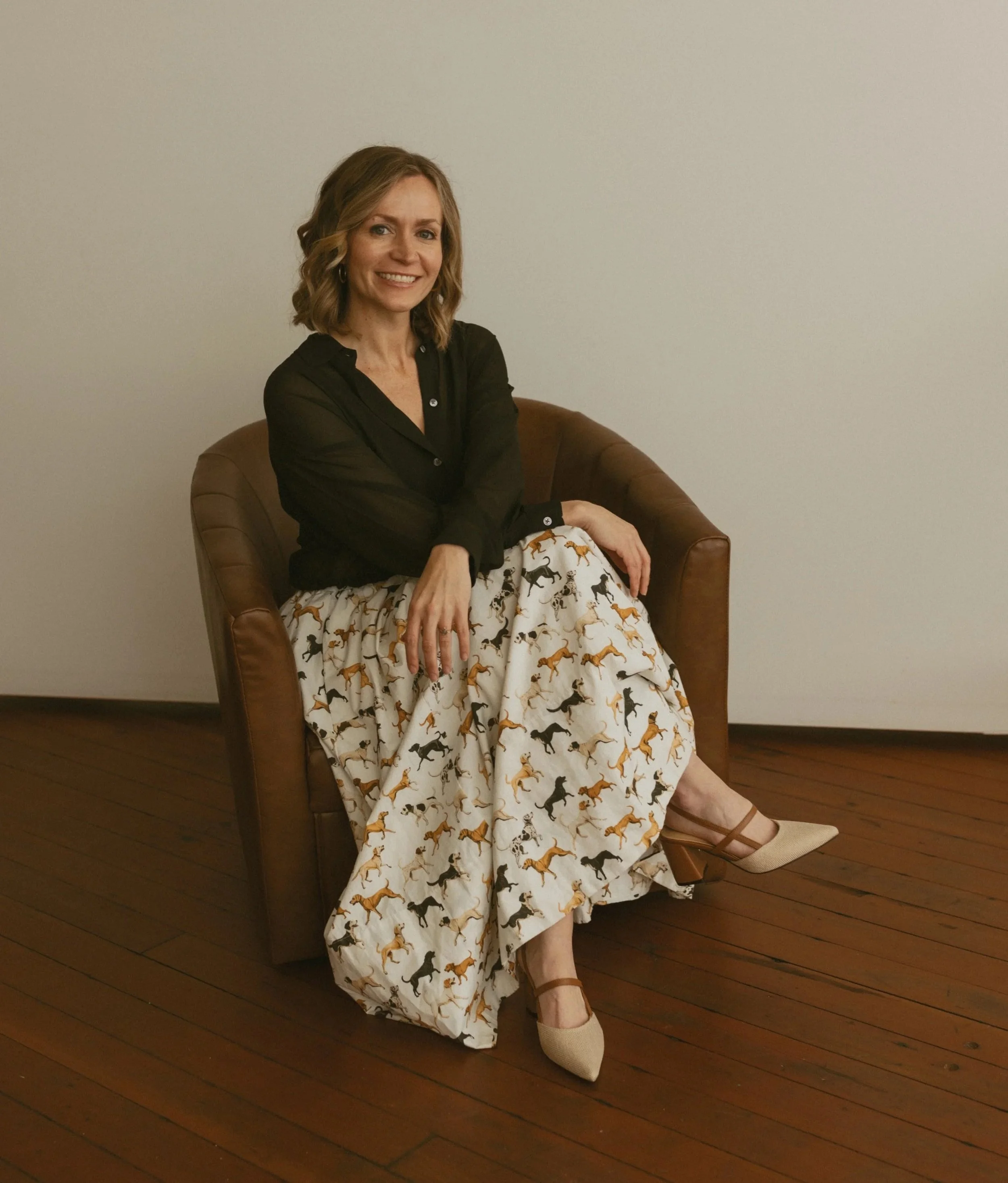 A woman sitting on a brown leather chair with a white dress featuring a pattern of various colored horses, and a black blouse, smiling, with her legs crossed, wearing beige shoes, in a room with wooden floor and plain off-white wall.