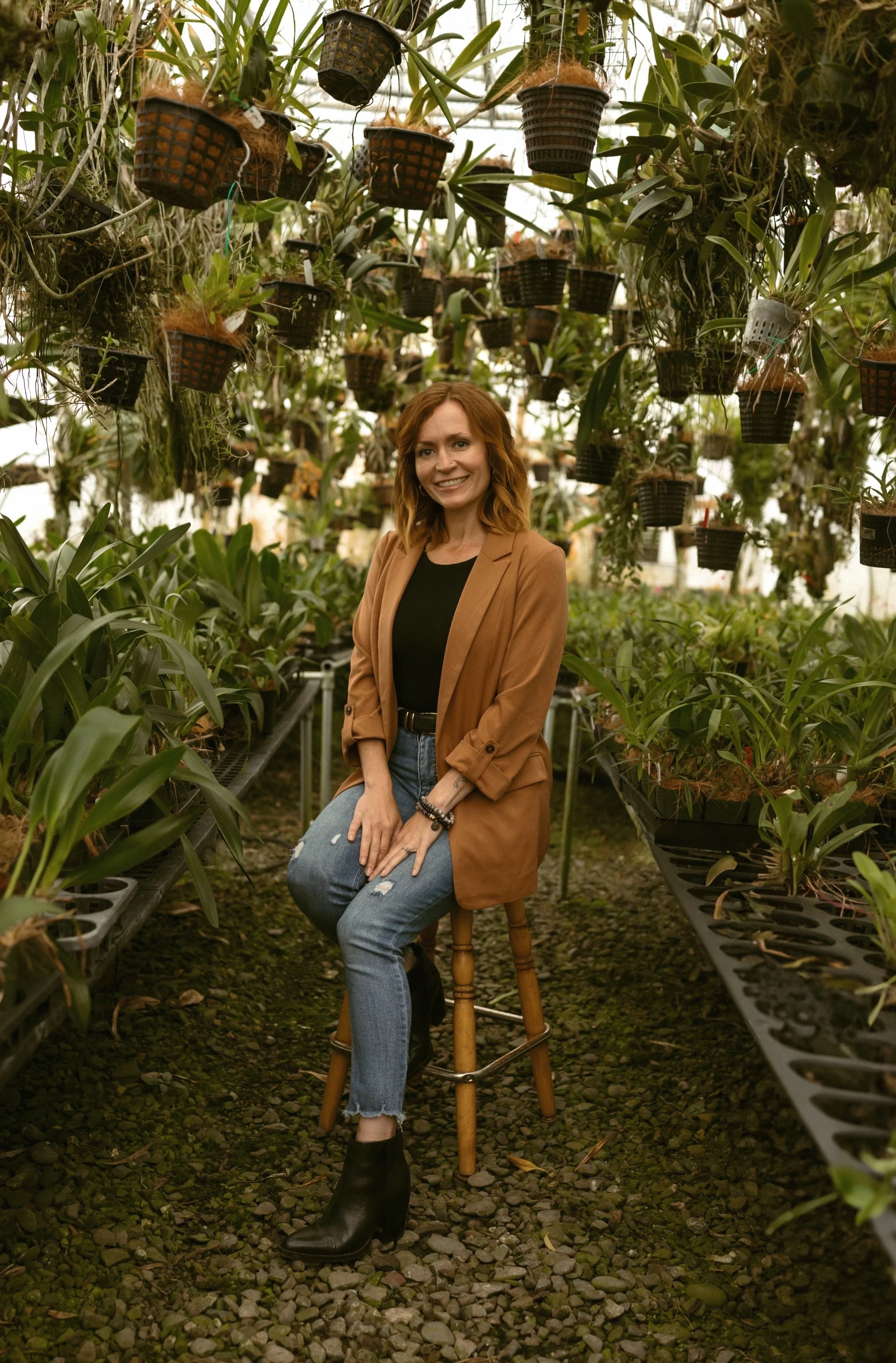 A smiling woman with red hair, wearing a tan blazer and ripped jeans, sitting on a wooden stool in a greenhouse filled with hanging potted plants.