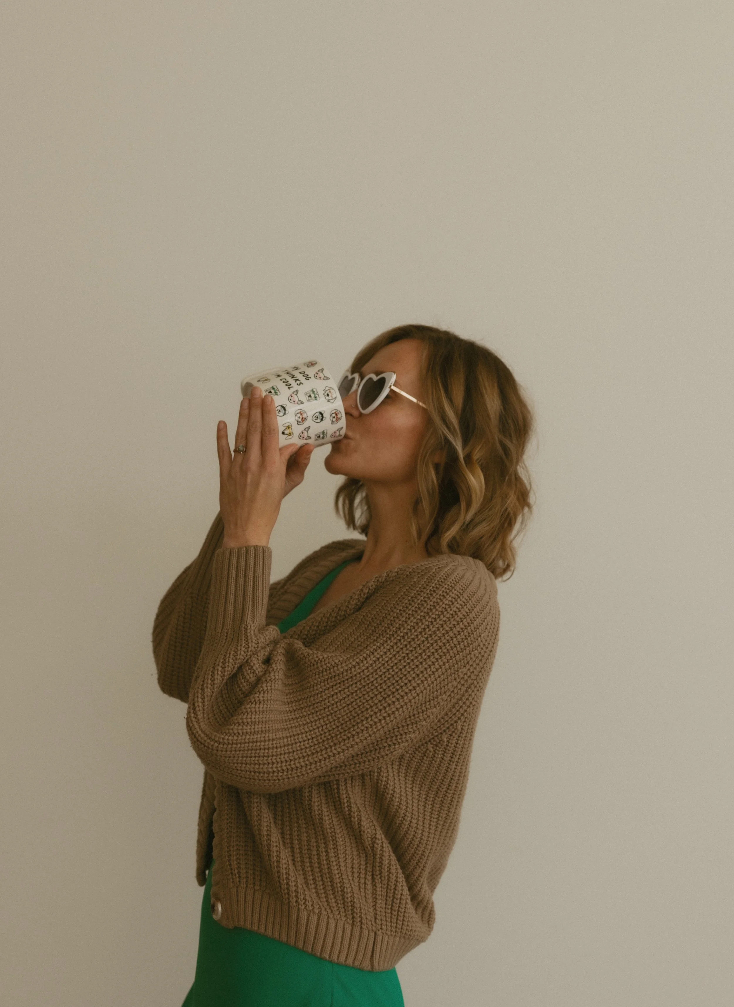 A woman with wavy light brown hair wearing heart-shaped sunglasses, a tan cardigan, and a green dress, drinking from a white mug with various small illustrations of animals and words on it, standing against a plain white wall.