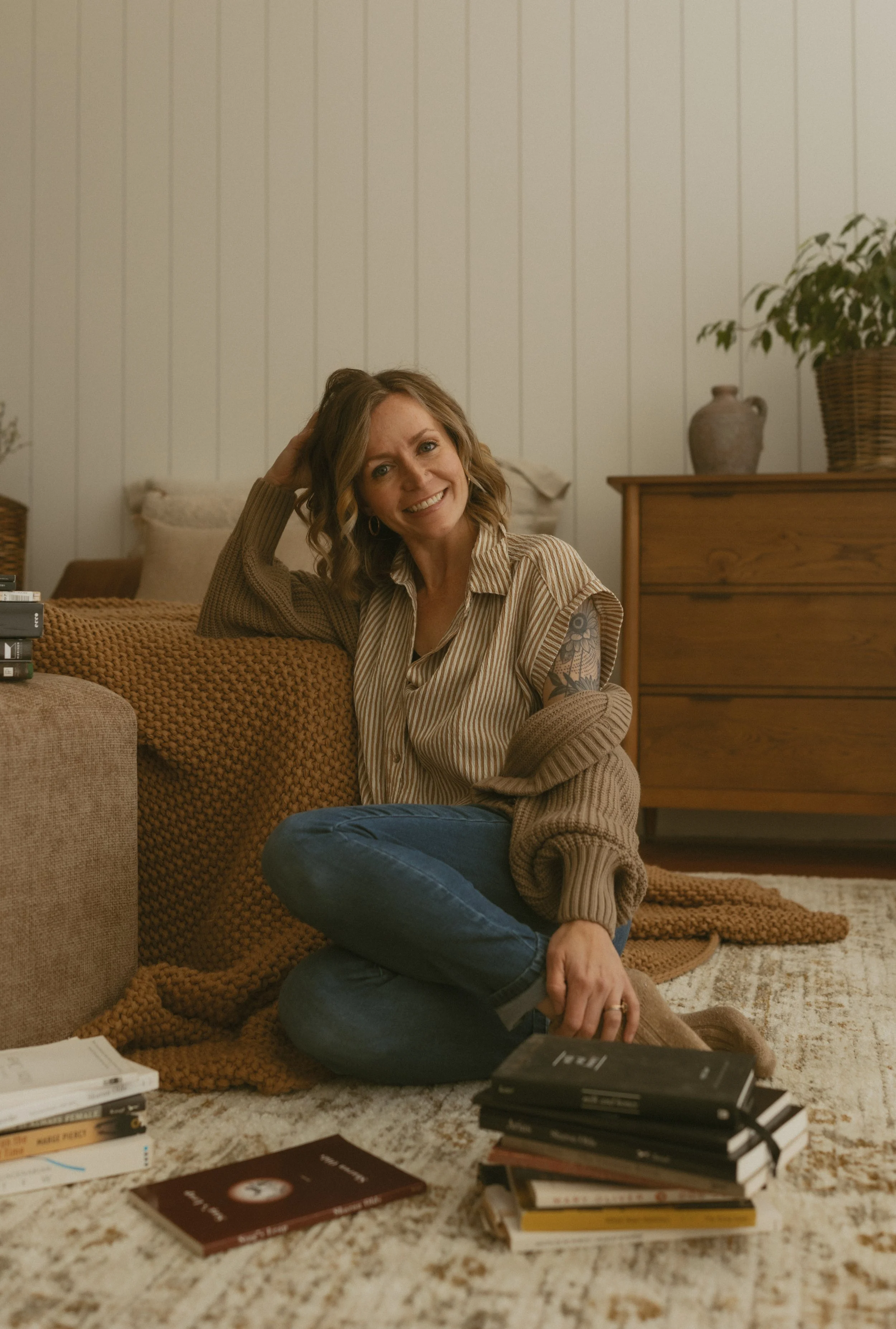 A woman with curly brown hair, tattoos on her arm, and wearing a beige striped shirt, is sitting on a carpeted floor in a cozy living room. She is smiling, with her head resting on her hand, surrounded by stacks of books, some open and some closed, and a beige knitted blanket. There is a wooden dresser with a brown vase and a potted plant in the background, with a white paneled wall behind her.