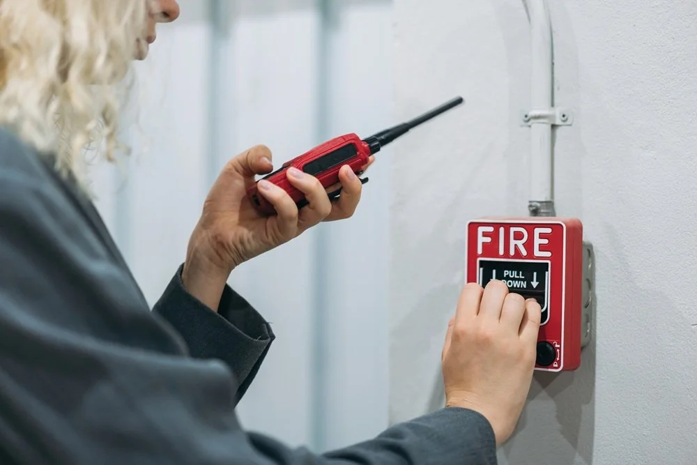 Woman in charcoal grey shirt with red walk talkie pulling Fire Alarm on wall