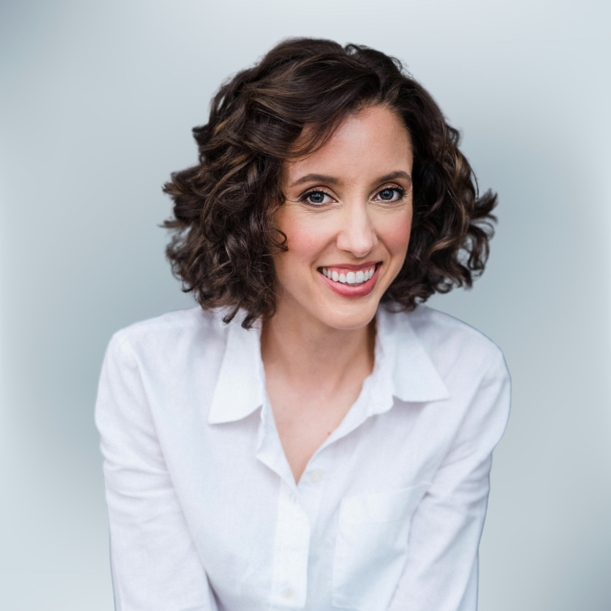 A woman with curly brown hair wearing a white shirt smiling at the camera.