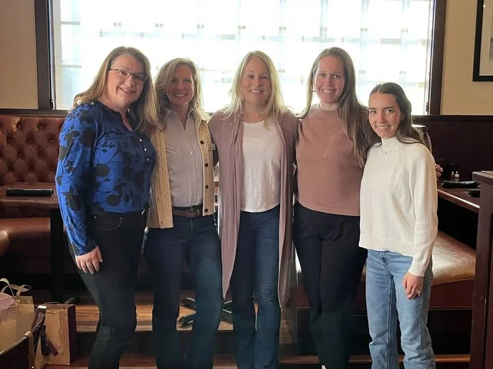Five women smiling and standing together inside a restaurant with booth seating and large windows in the background.