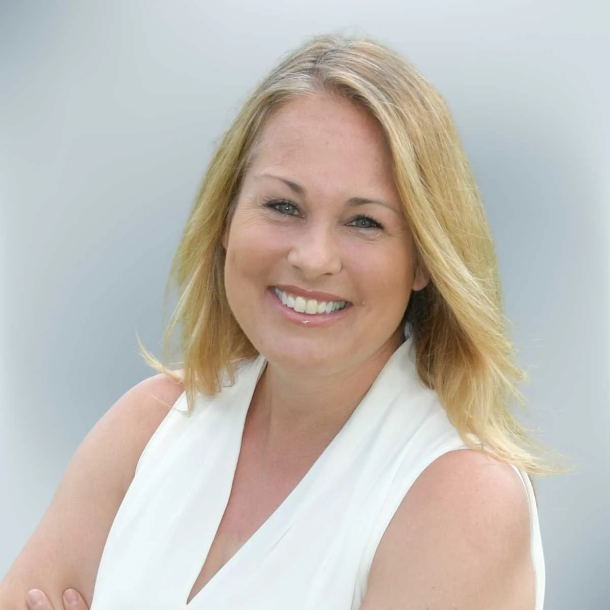 A smiling woman with blonde hair wearing a sleeveless white top against a light gray background.