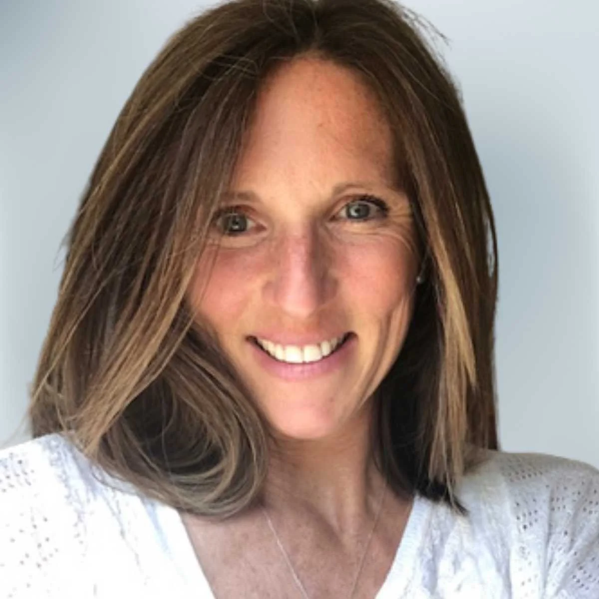 Close-up of a smiling woman with shoulder-length brown hair, wearing a white top, against a light gray background.