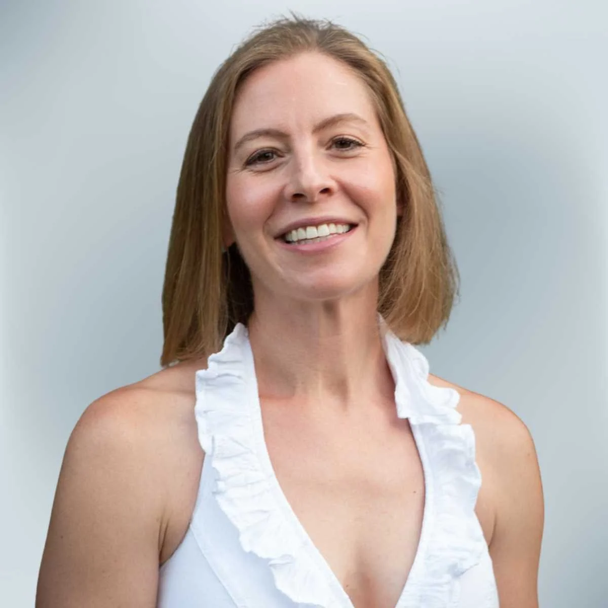 A woman with shoulder-length red hair smiling at the camera, wearing a white sleeveless top with ruffles, against a light gray background.