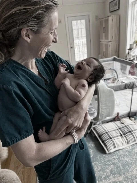 A woman holding a smiling newborn baby in a living room with a dog crate and a patterned pillow in the background.
