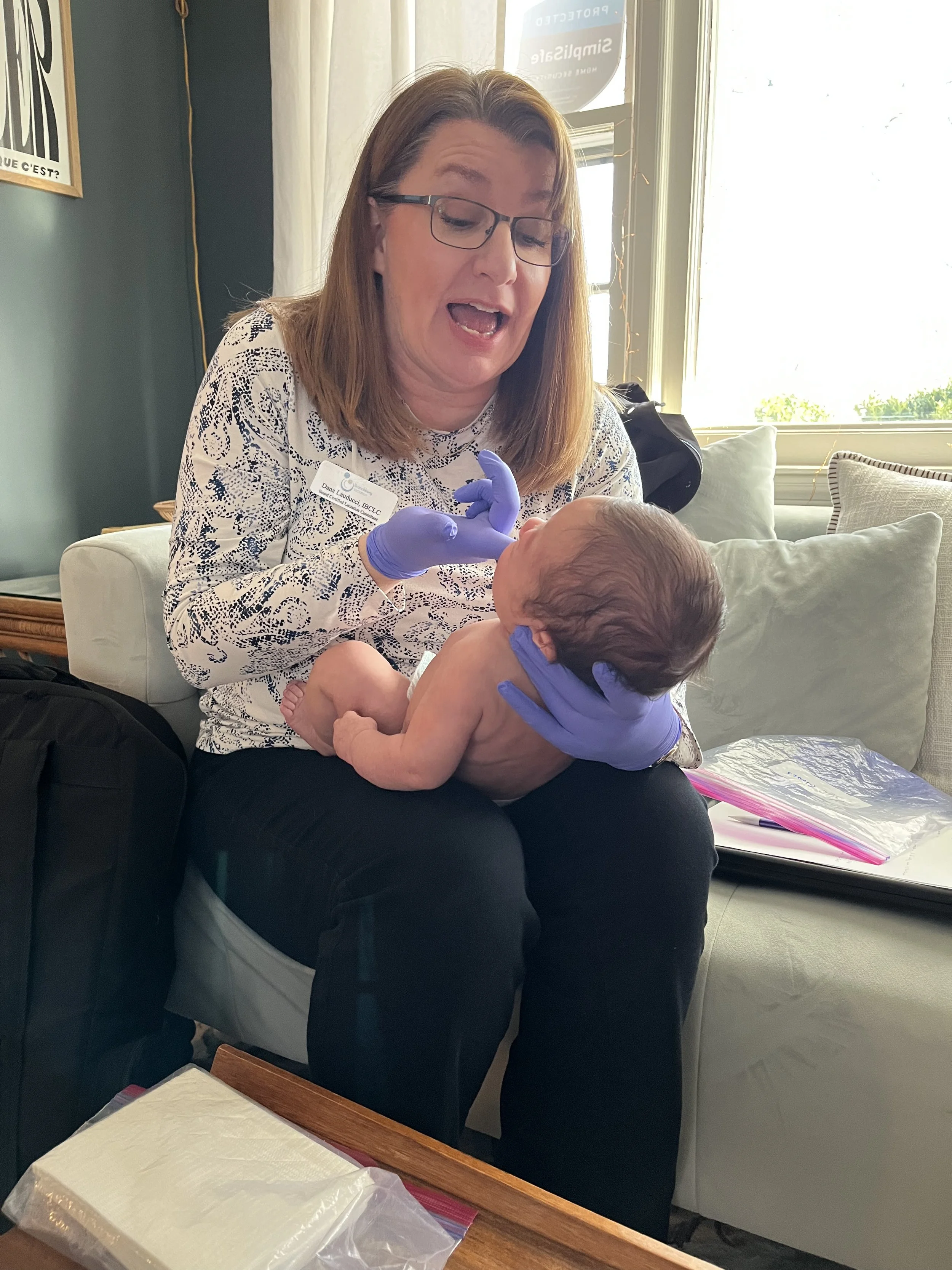 A woman with glasses and purple gloves holding a newborn baby in a room with sunlight coming through the window, sitting on a beige sofa with papers and a plastic bag nearby.