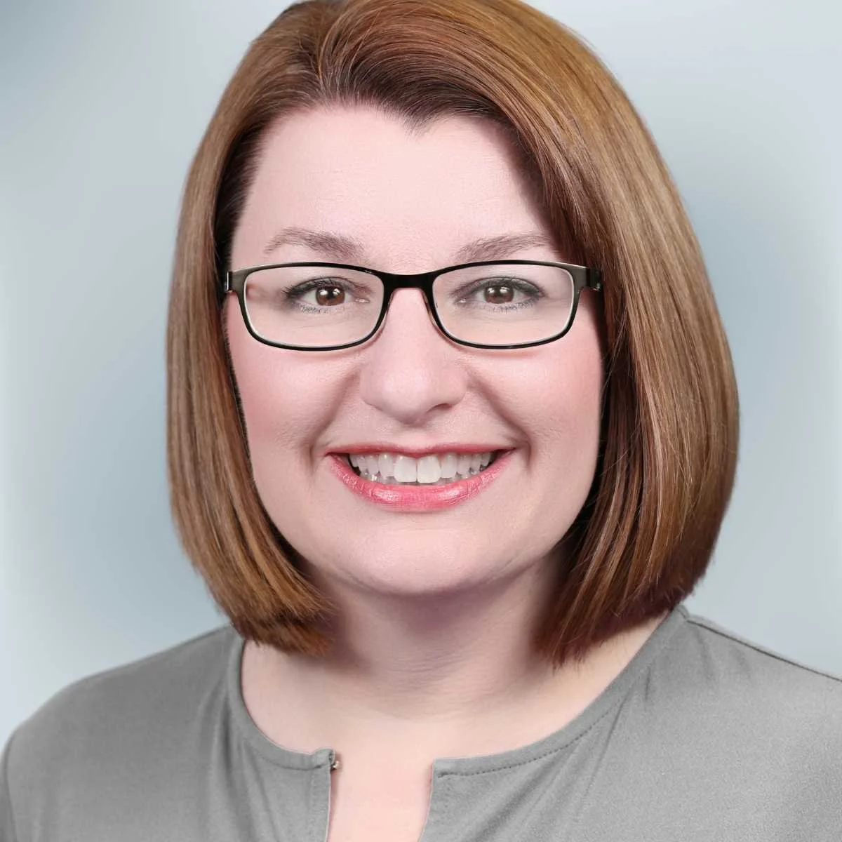 A woman with shoulder-length reddish-brown hair, glasses, and a gray top, smiling at the camera against a plain light background.