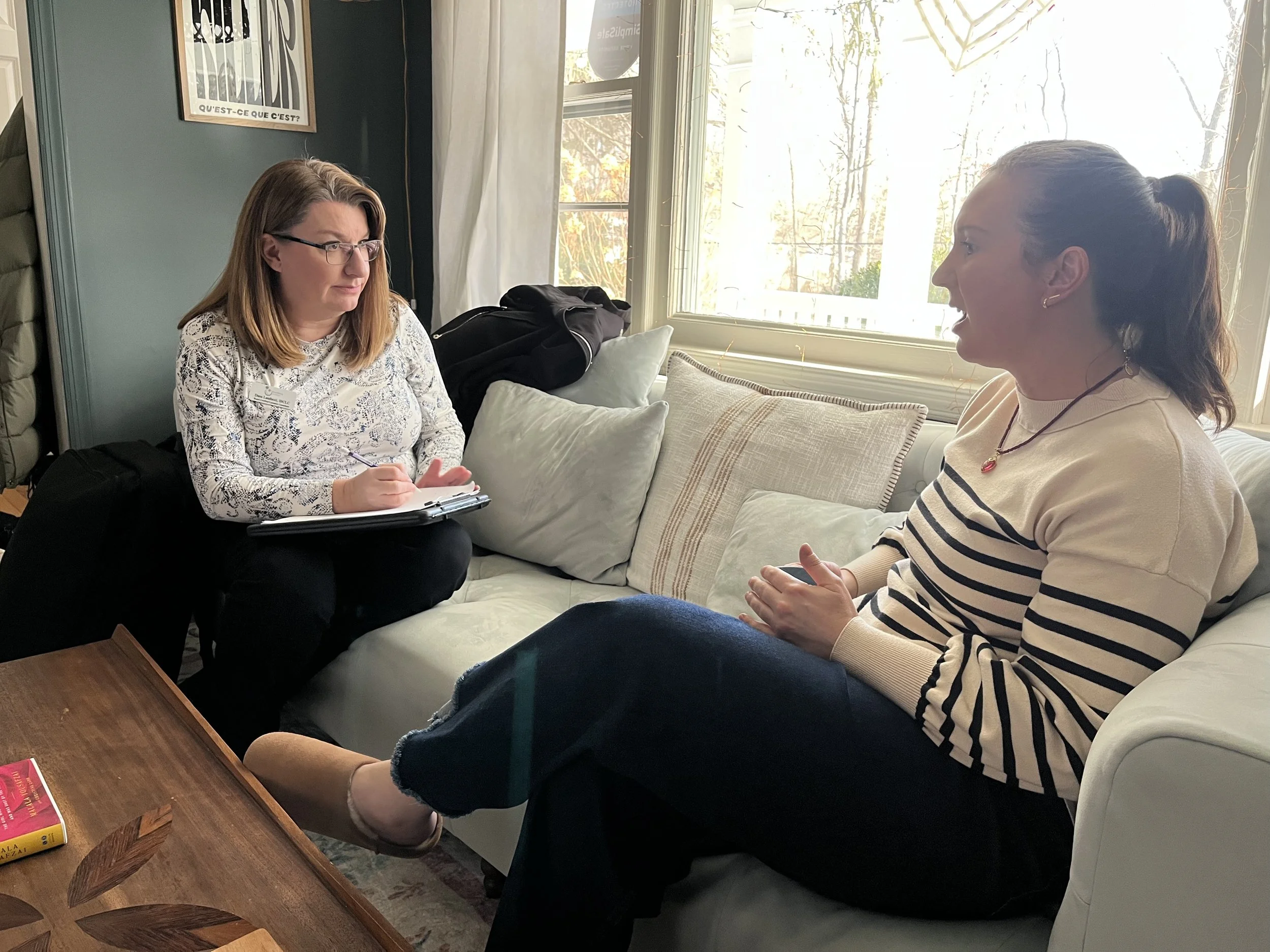 Two women sitting and having a conversation in a cozy living room. One woman is taking notes, the other is speaking animatedly.