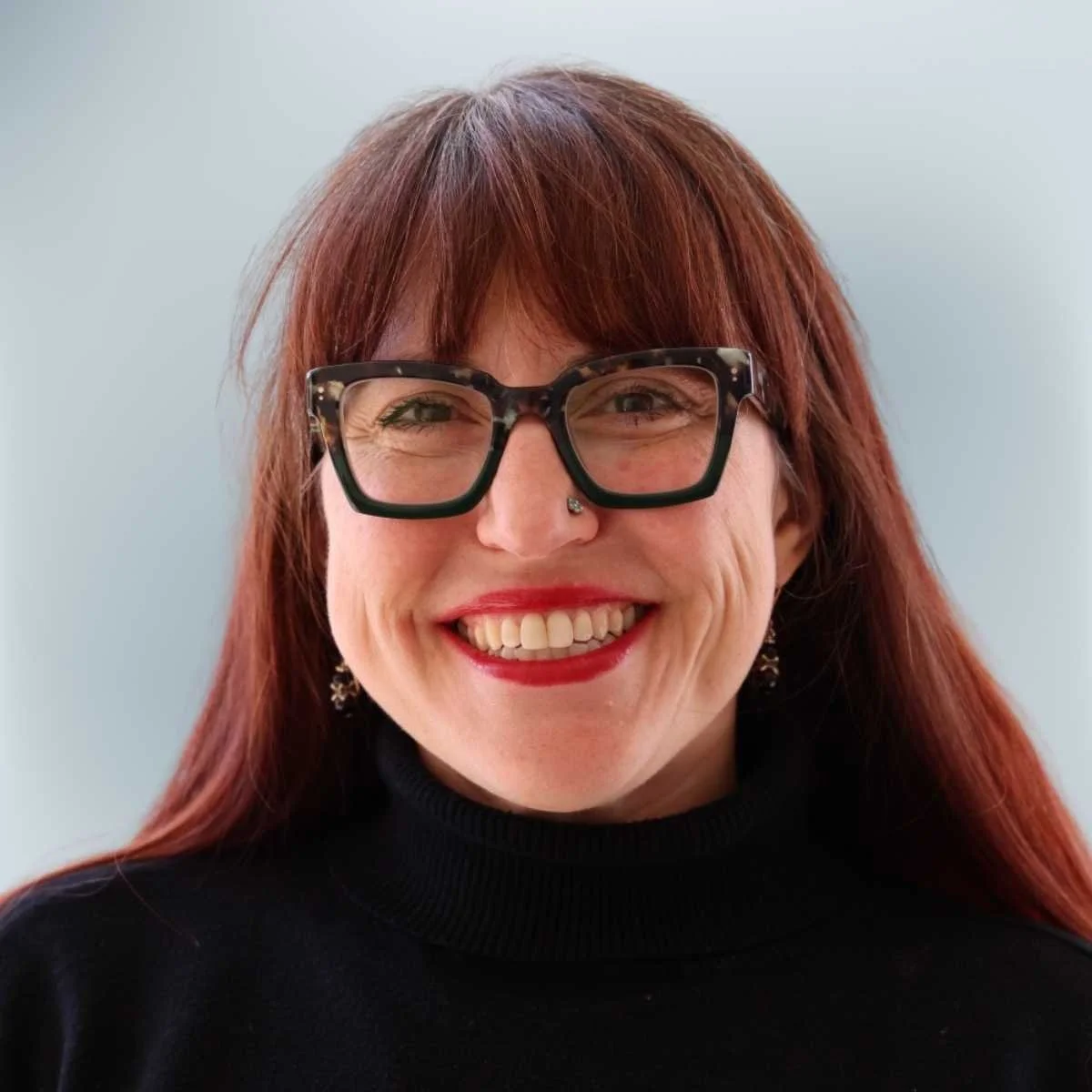 A woman with red hair, wearing large black glasses with a tortoiseshell pattern, smiling. She is dressed in a black top and has earrings and a nose stud.