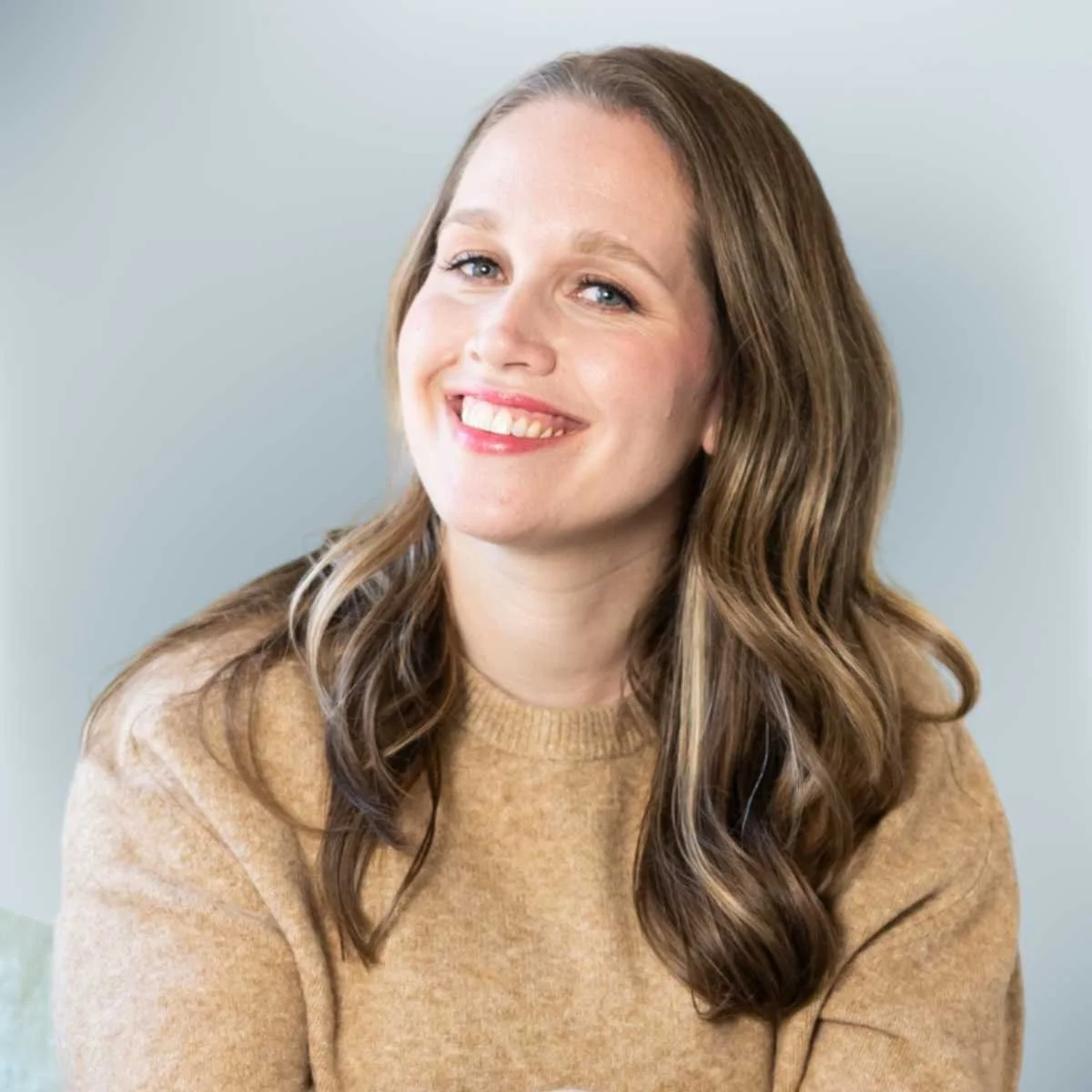 A smiling woman with wavy brown hair wearing a beige sweater.