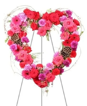 A heart-shaped floral arrangement made of pink and red roses, pink carnations, purple flowers, and decorative brown elements, supported by a metal stand.