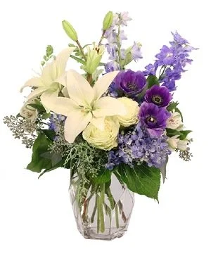 A floral arrangement with white lilies, purple anemones, purple and blue delphiniums, and other mixed flowers in a clear glass vase.