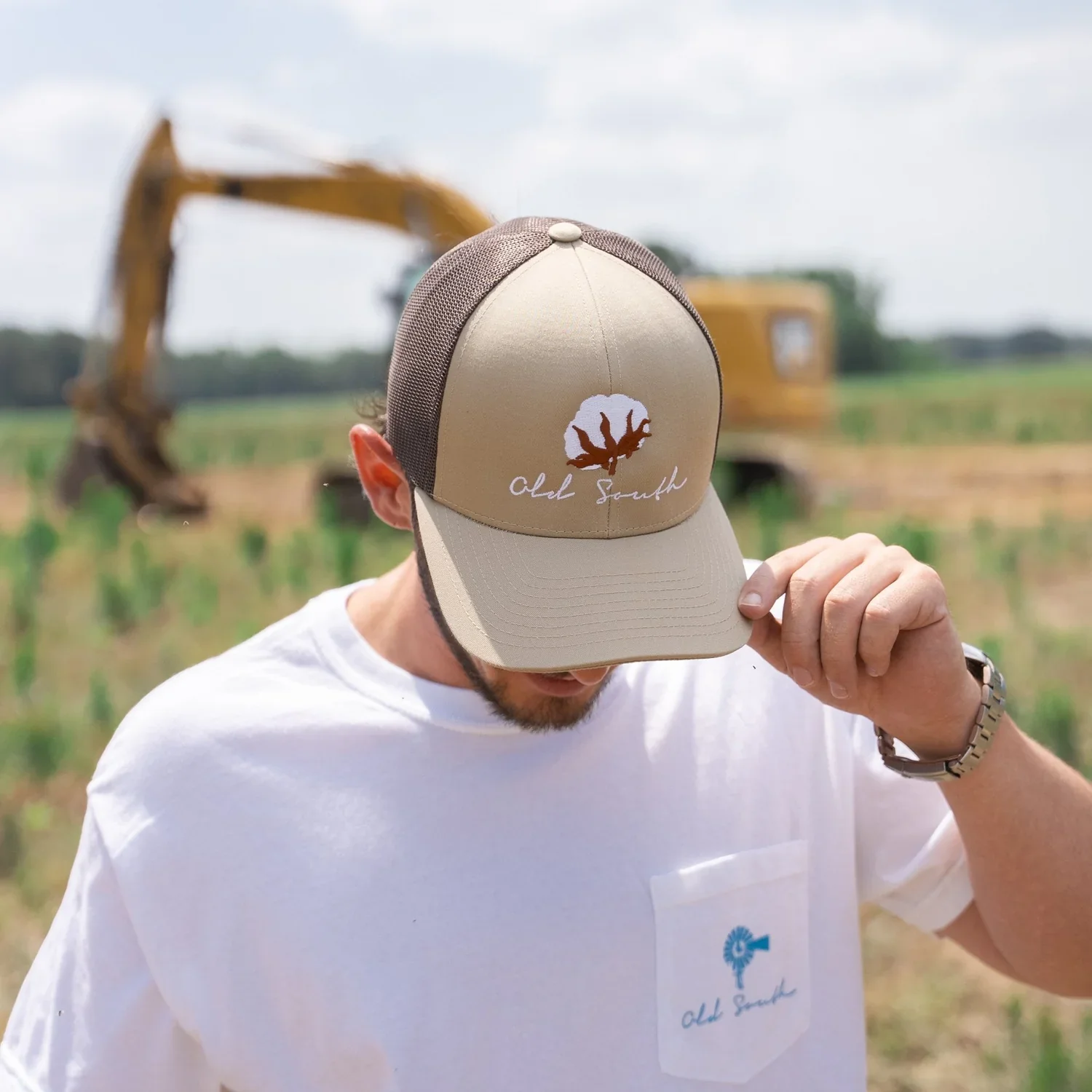 A man wearing a white T-shirt and a beige trucker hat with 'Old South' and a tree logo in front of a yellow construction excavator in a field.