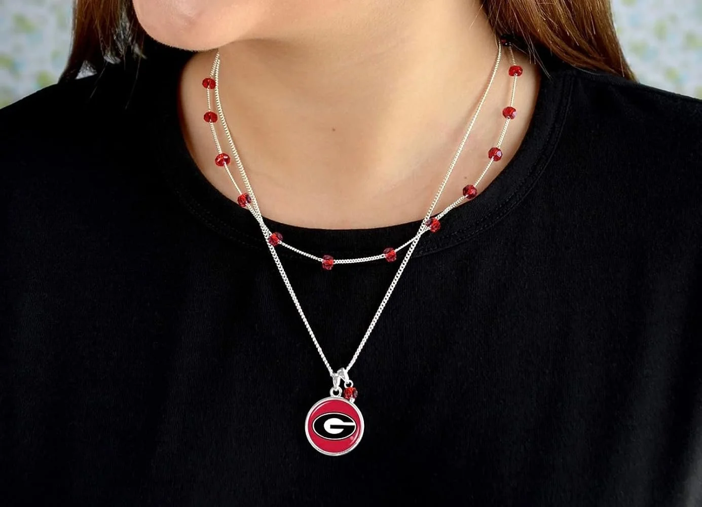 A person is wearing two layered necklaces, one with red beads and the other with a University of Georgia logo pendant, all against a black shirt.