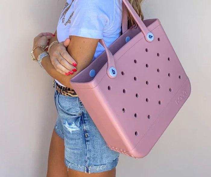 A woman wearing a blue t-shirt and distressed denim shorts carries a large pink tote bag with circular holes and blue buttons, standing against a neutral background.