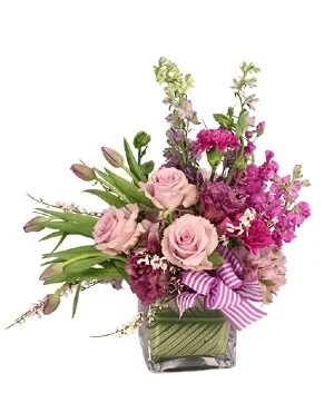 A floral arrangement with pink roses, purple alstroemeria, and various greenery in a square glass vase, decorated with a purple and white striped ribbon.
