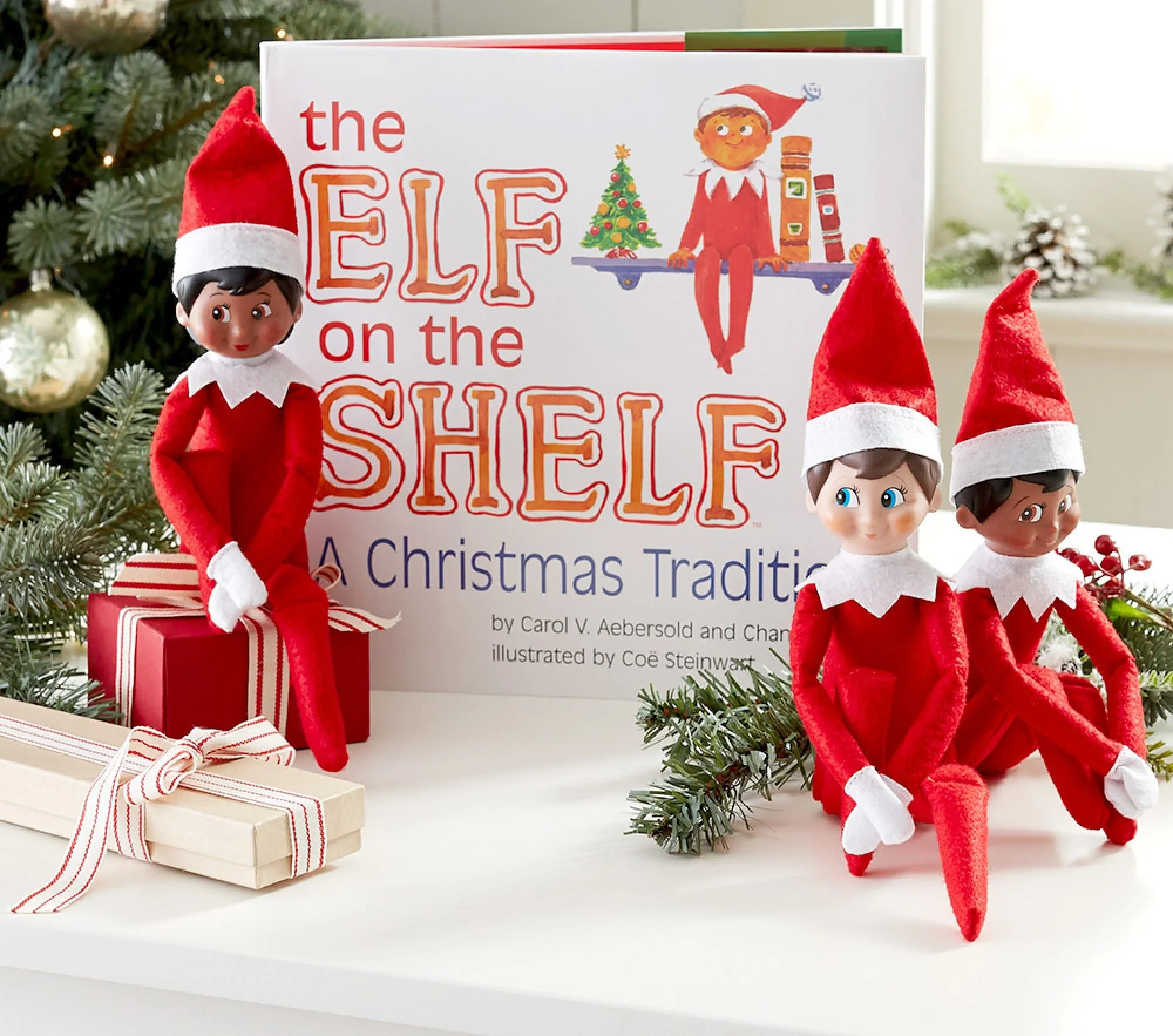 Decorative Christmas scene featuring three elf dolls in red outfits with white collars and red hats, a Christmas book titled 'The Elf on the Shelf', with a Christmas tree illustration, and wrapped gifts with ribbons, surrounded by pine branches and ornaments.