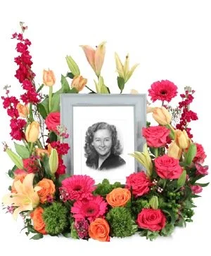 A framed black and white photo of a smiling woman surrounded by a colorful floral arrangement of pink, orange, yellow, and peach flowers.
