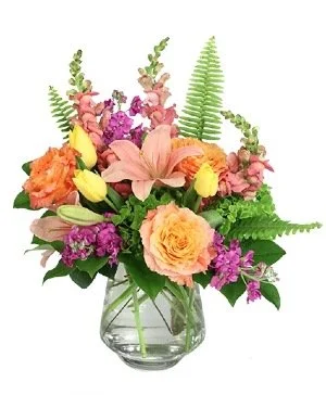 Colorful flower bouquet in a clear glass vase, featuring pink, orange, yellow, and purple flowers with green foliage.