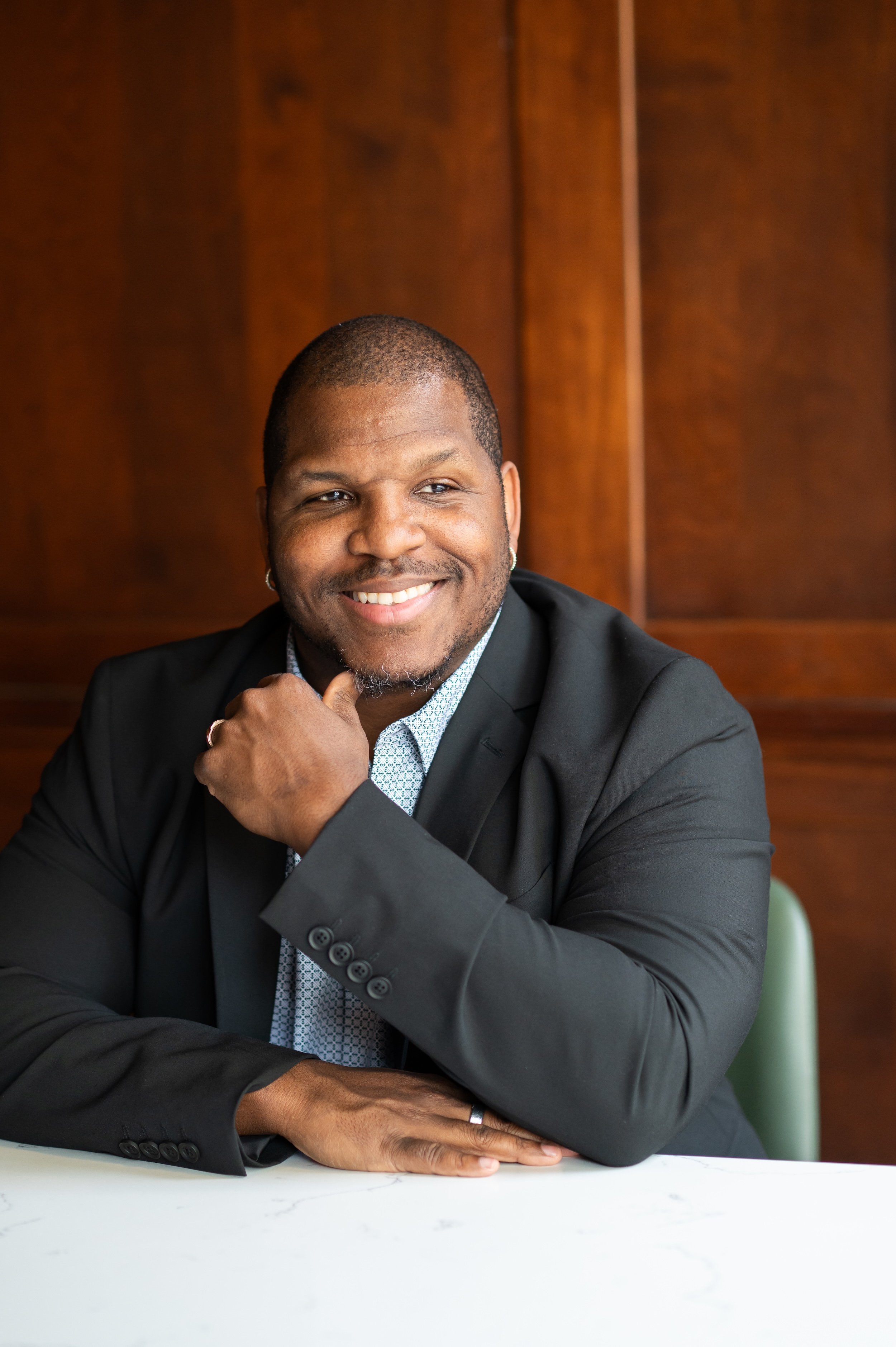 Smiling man sitting at a white table with a wooden background, wearing a black blazer and light-patterned shirt.
