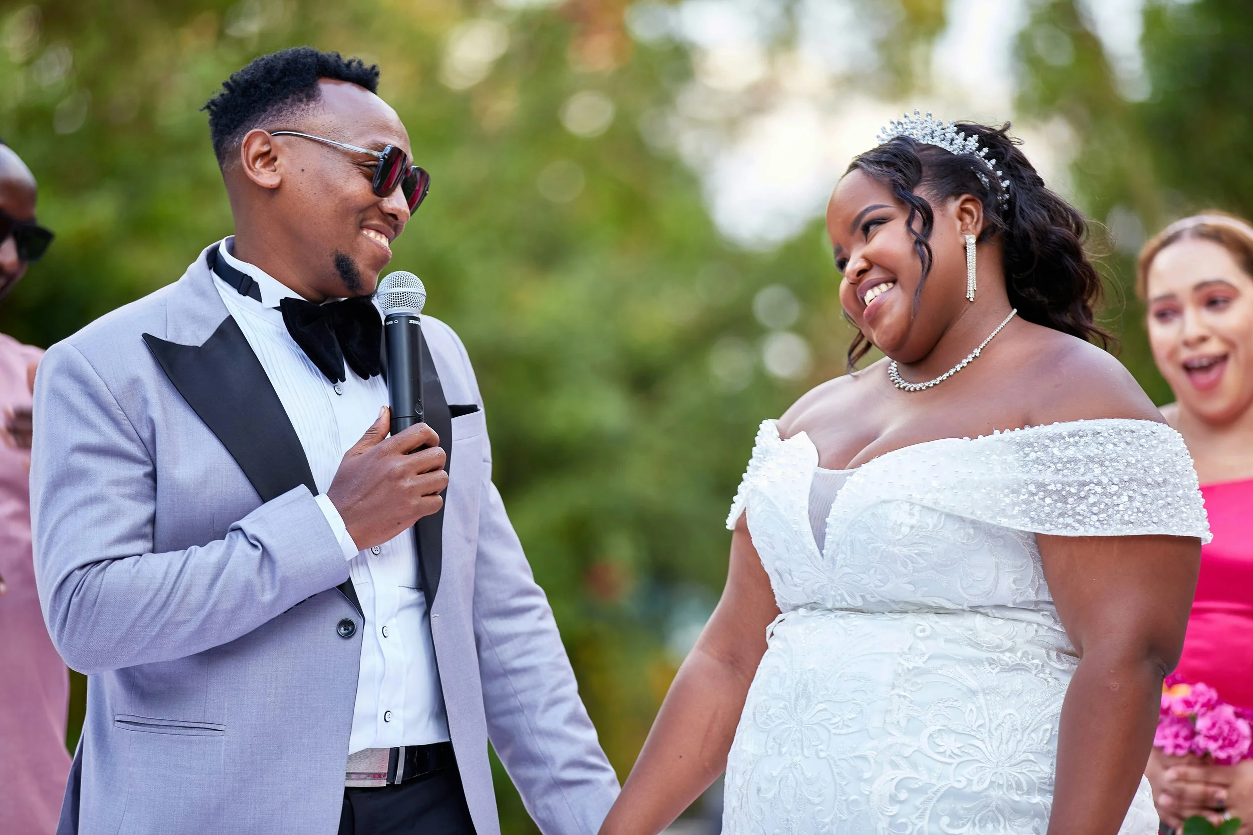A wedding ceremony outdoors with a groom in a tuxedo holding a microphone and a bride in a white wedding dress wearing a tiara, smiling at each other.